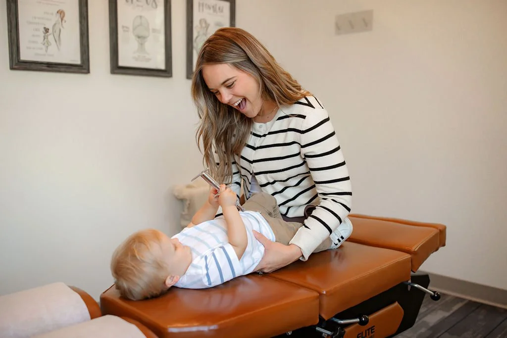 A young woman and a small child lying on a medical examination table in a doctor's office, smiling and playing with a stethoscope.