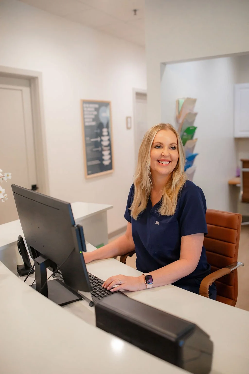 A smiling woman with blonde hair, wearing a navy blue uniform and smartwatch, sitting at a reception desk with a computer in a modern office or clinic environment.