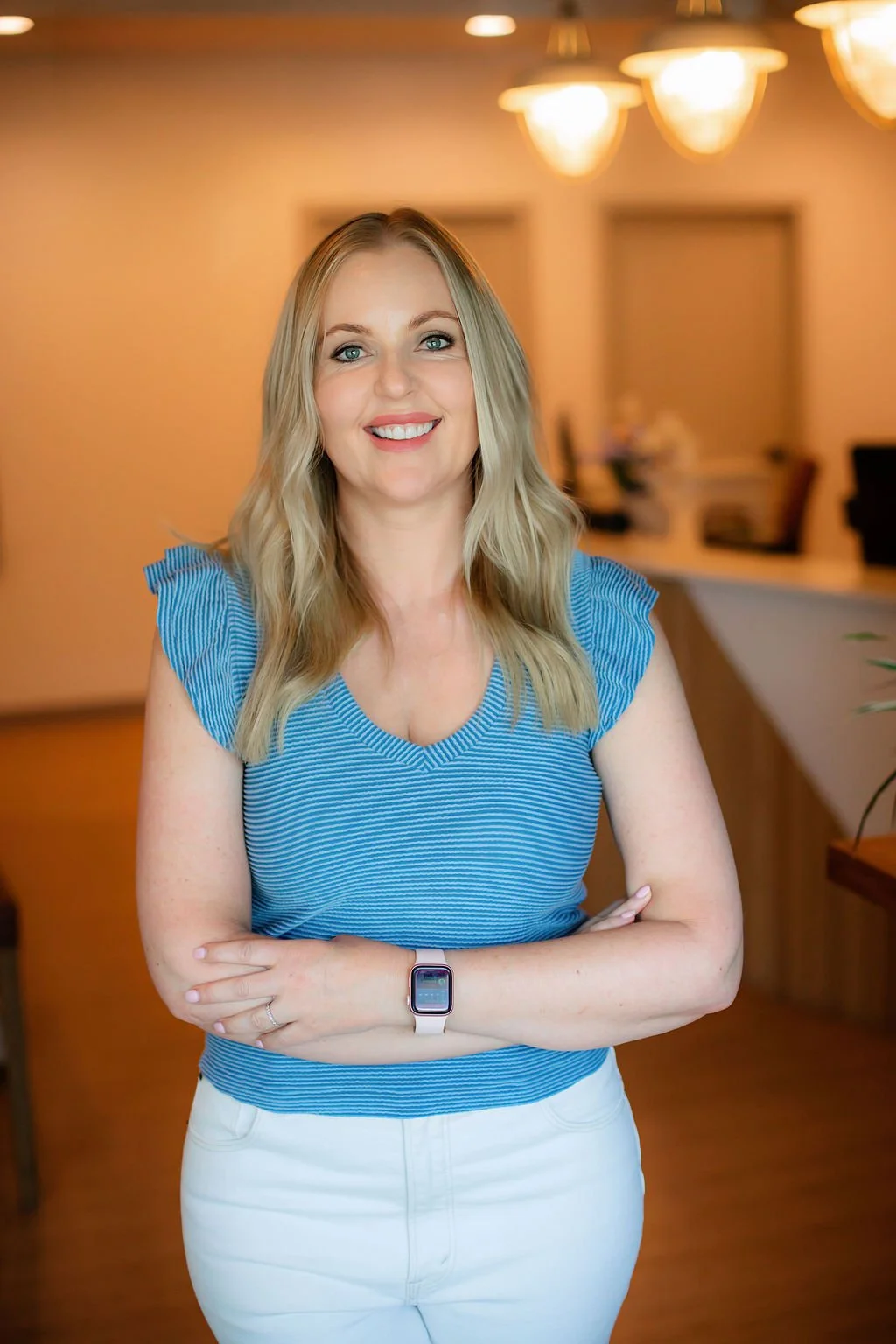 A smiling woman with blonde hair wearing a blue striped top and white pants standing in an indoor setting with warm lighting.