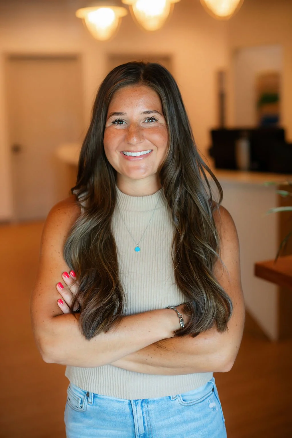 A smiling woman with long brown hair and freckles, wearing a sleeveless beige turtleneck top, blue jeans, a necklace with a small blue pendant, and a bracelet, standing indoors with a blurred background of warm lighting and furniture.