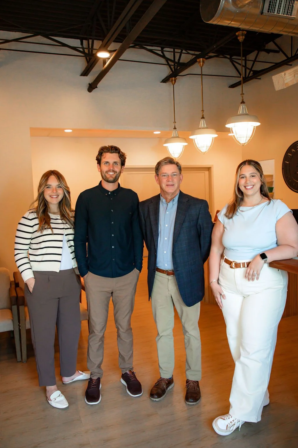 Four smiling people standing indoors in a modern room with pendant lights and a wall clock. From left to right: a young woman in a striped shirt and brown pants, a young man in a black shirt and beige pants, an older man in a blazer and khakis, and a young woman in a white top and pants.
