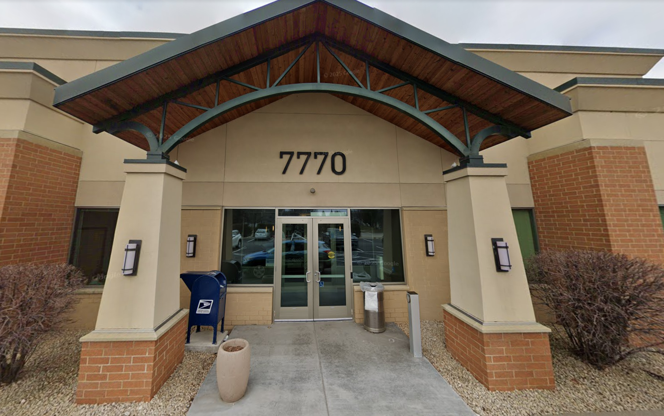 Front entrance of a commercial building with the number 7770 above the door, glass double doors, mailbox, trash can, and bushes on either side of the entrance.
