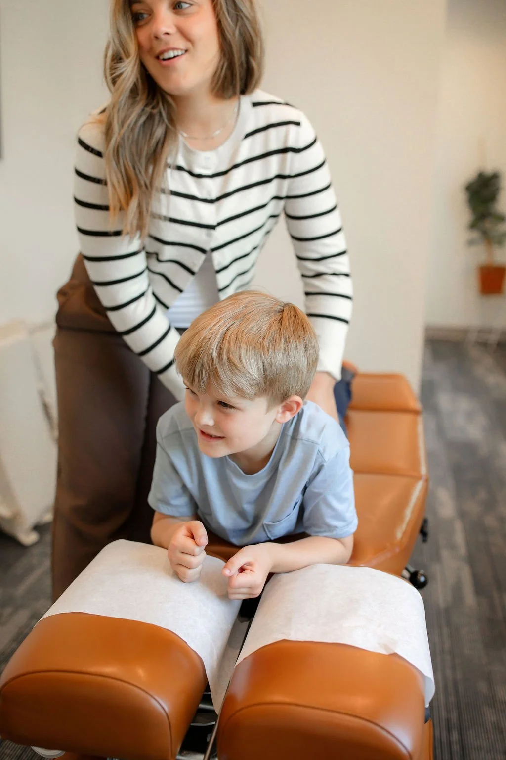 A woman and a young boy in a therapy or chiropractic setting. The boy is lying face down on a padded table with his fists clenched, smiling, while the woman, standing behind him, appears to be giving instruction or encouragement. The woman is wearing a striped shirt, and the boy is wearing a light blue t-shirt.