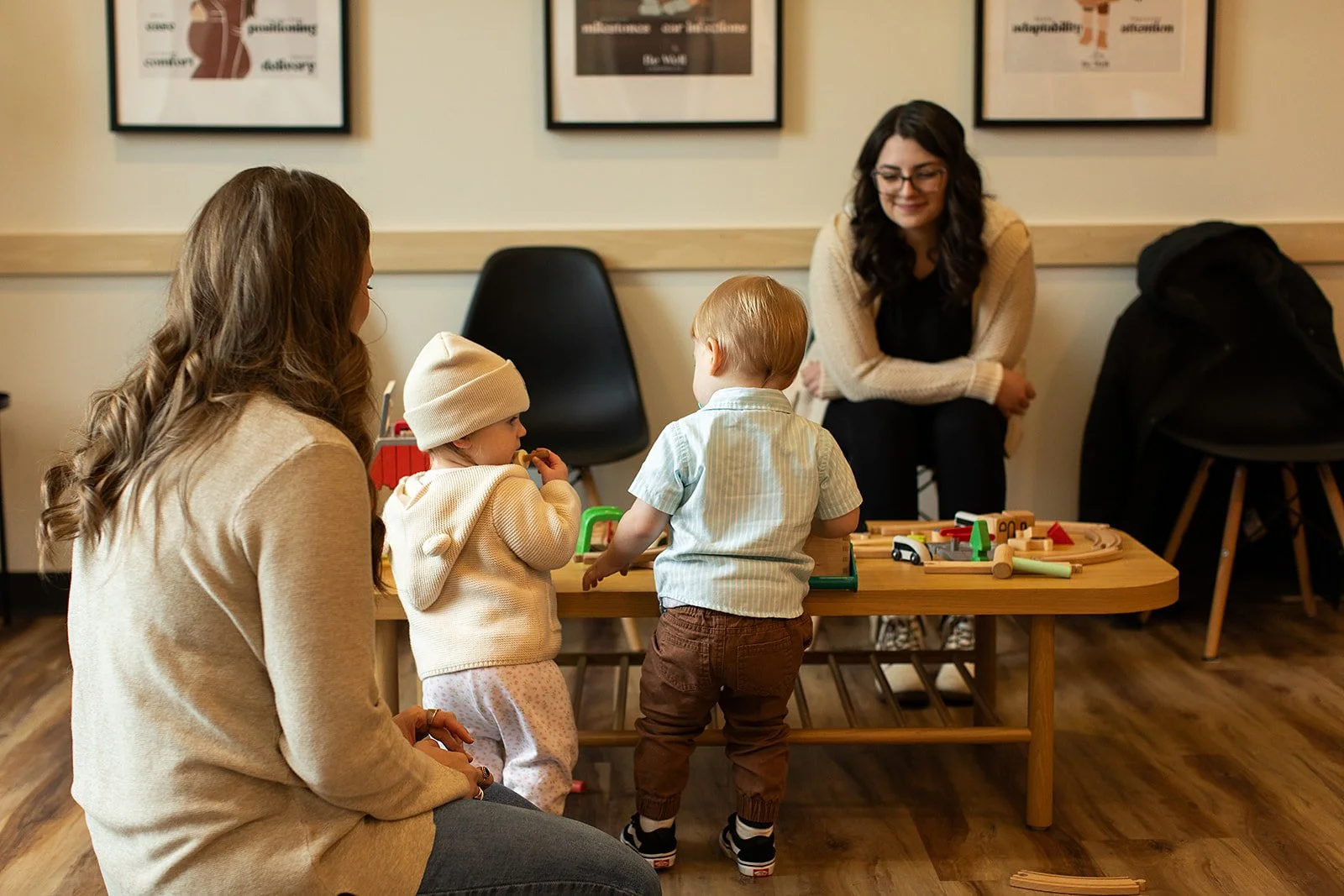 Two women and three children playing with wooden toys at a table in a cozy room with framed pictures on the wall.