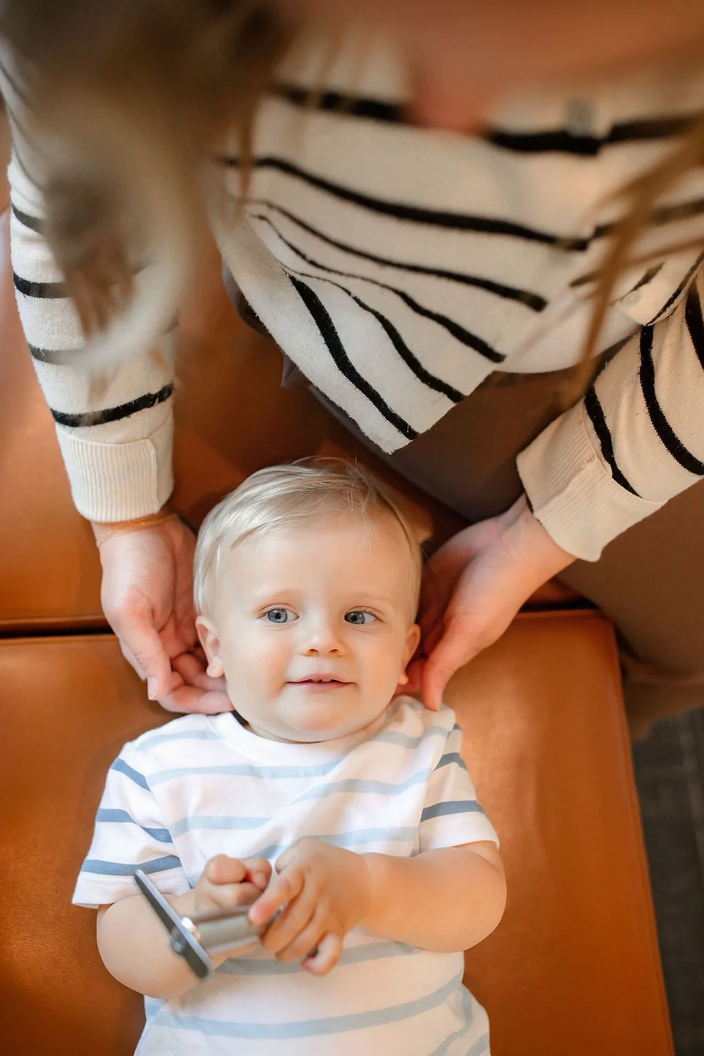 A young boy with blond hair and blue eyes lying on a leather couch, looking upward, holding a remote control. An adult, likely a woman, is sitting above him, wearing a beige sweater with black stripes, and is gently holding his head with her hands.