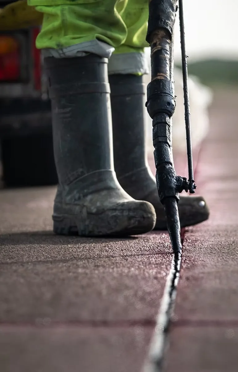 Close-up of a worker in rubber boots and high-visibility clothing applying black waterproof coating along a seam on a concrete surface using a caulking gun.