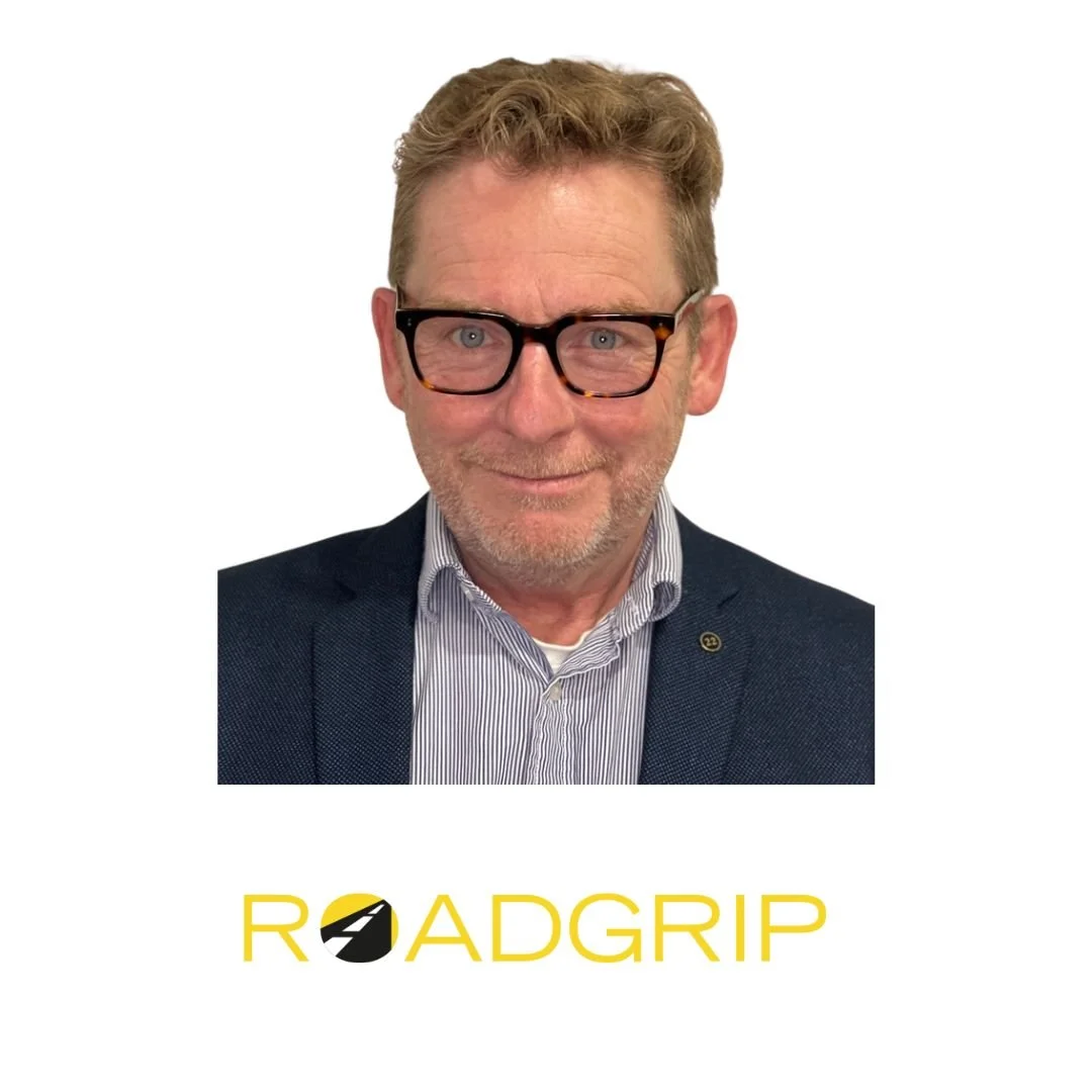 A middle-aged man with light brown hair, glasses, and a short beard, smiling at the camera, wearing a dark blazer and a striped dress shirt, with the text 'ROADGRIP' beneath him that features a stylized road icon.