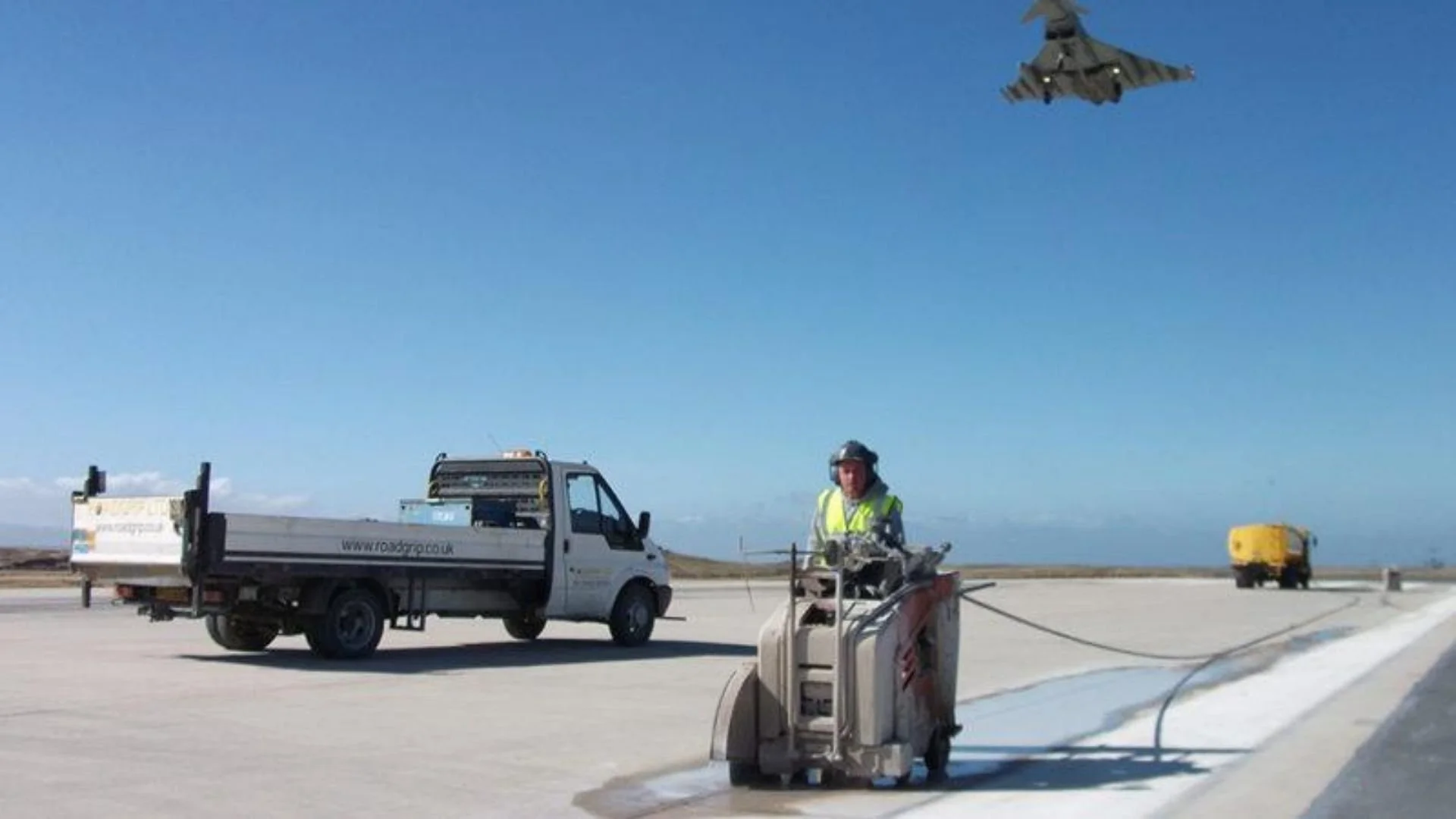 A worker with safety gear operating machinery on a runway or tarmac, with a small truck and distant aircraft in the background, under a clear blue sky.