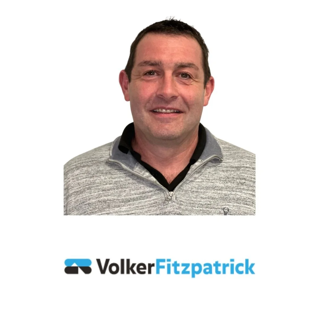 Photograph of a man with short dark hair, wearing a light gray zip-up sweater over a black collared shirt, smiling at the camera, with the name 'Volker Fitzpatrick' and logo below.