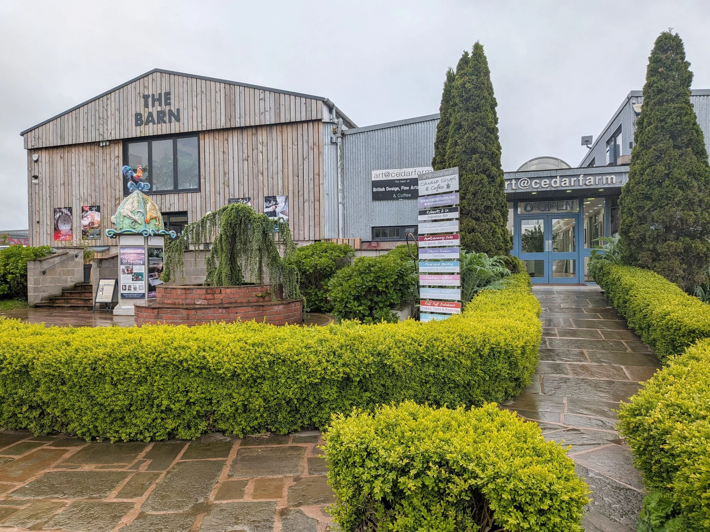 Exterior of a building at Cedar Farm in Mawdesley with wooden and metal panels, called 'The Barn'. There are two tall evergreen trees flanking the entrance, which has glass doors with an 'Open' sign. Decorative plants and hedging with a sign listing 