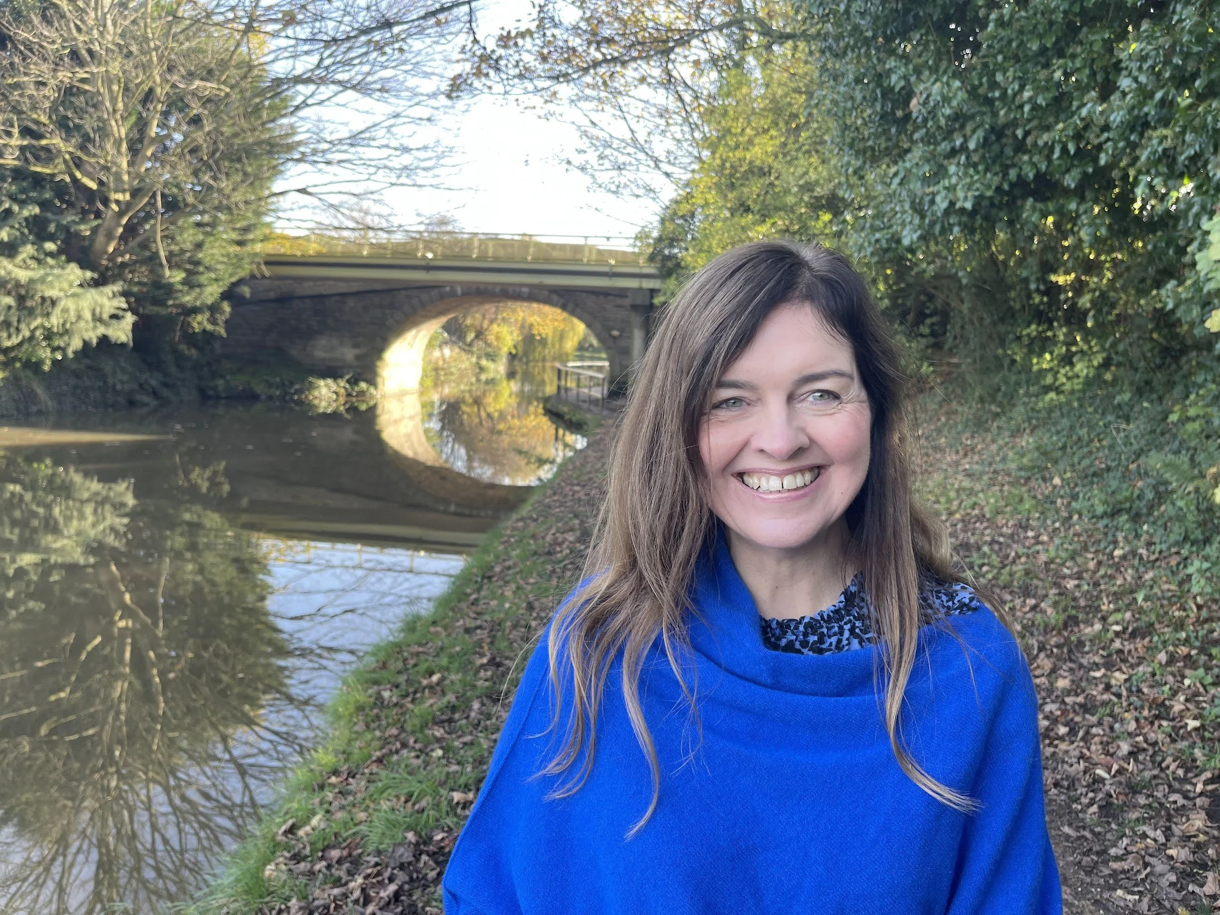 A picture of therapist, Davina Hanlon, a smiling woman with long brown hair and green eyes taking a selfie outdoors by the Leeds Liverpool canal, with trees and a stone bridge in the background.