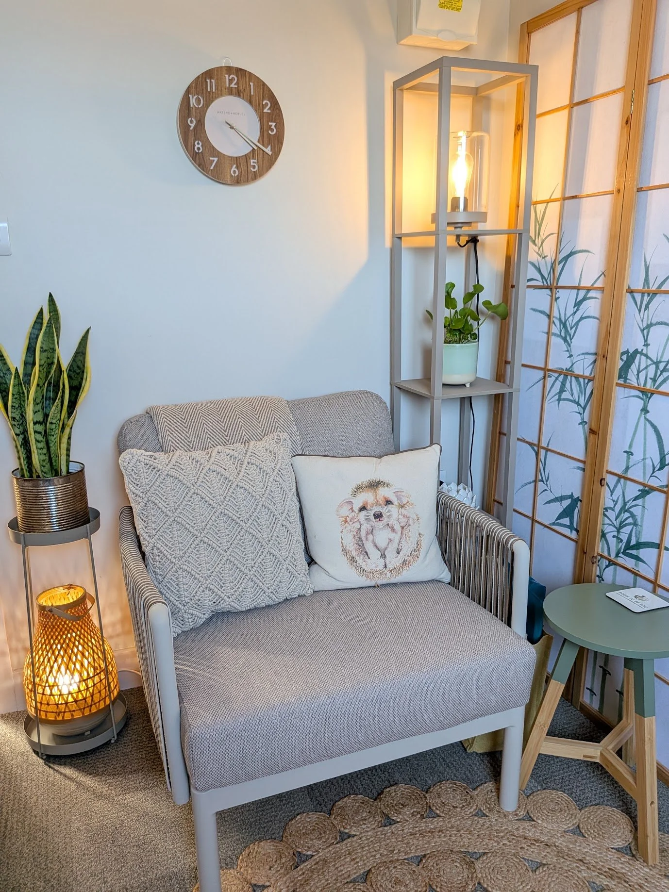 A chair in a therapy room  that has a cushion with a hedgehog illustration, a small round sage-green side table, a decorative wicker floor lamp, a tall grey shelving unit with a potted plant, a wooden room divider with a bamboo design, a wall clock w