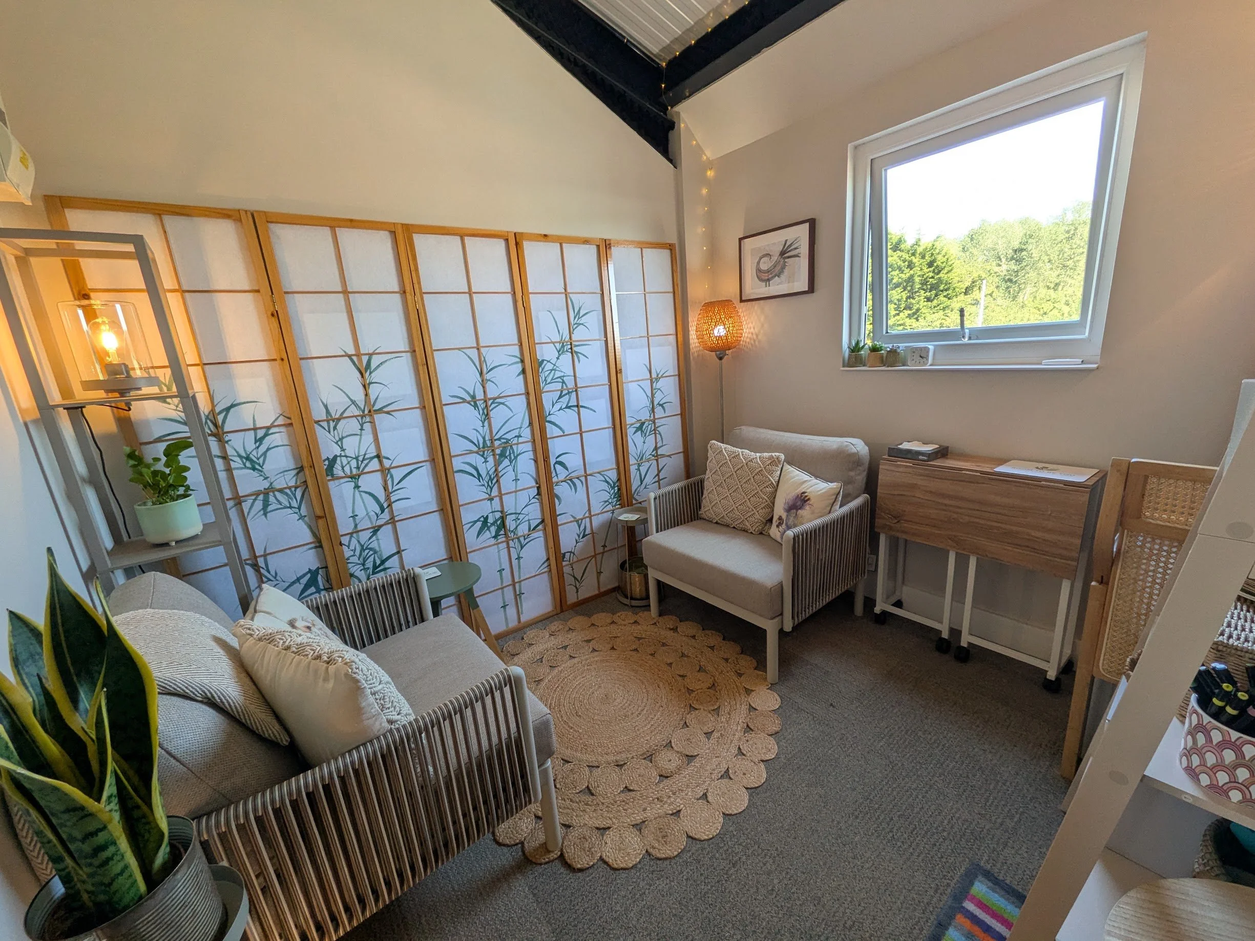 Cosy therapy room with comfortable chairs, a small side table, a wooden room divider with a bamboo design, a floor lamp with a woven shade, a window showing greenery outside, and various plants and decorative items.