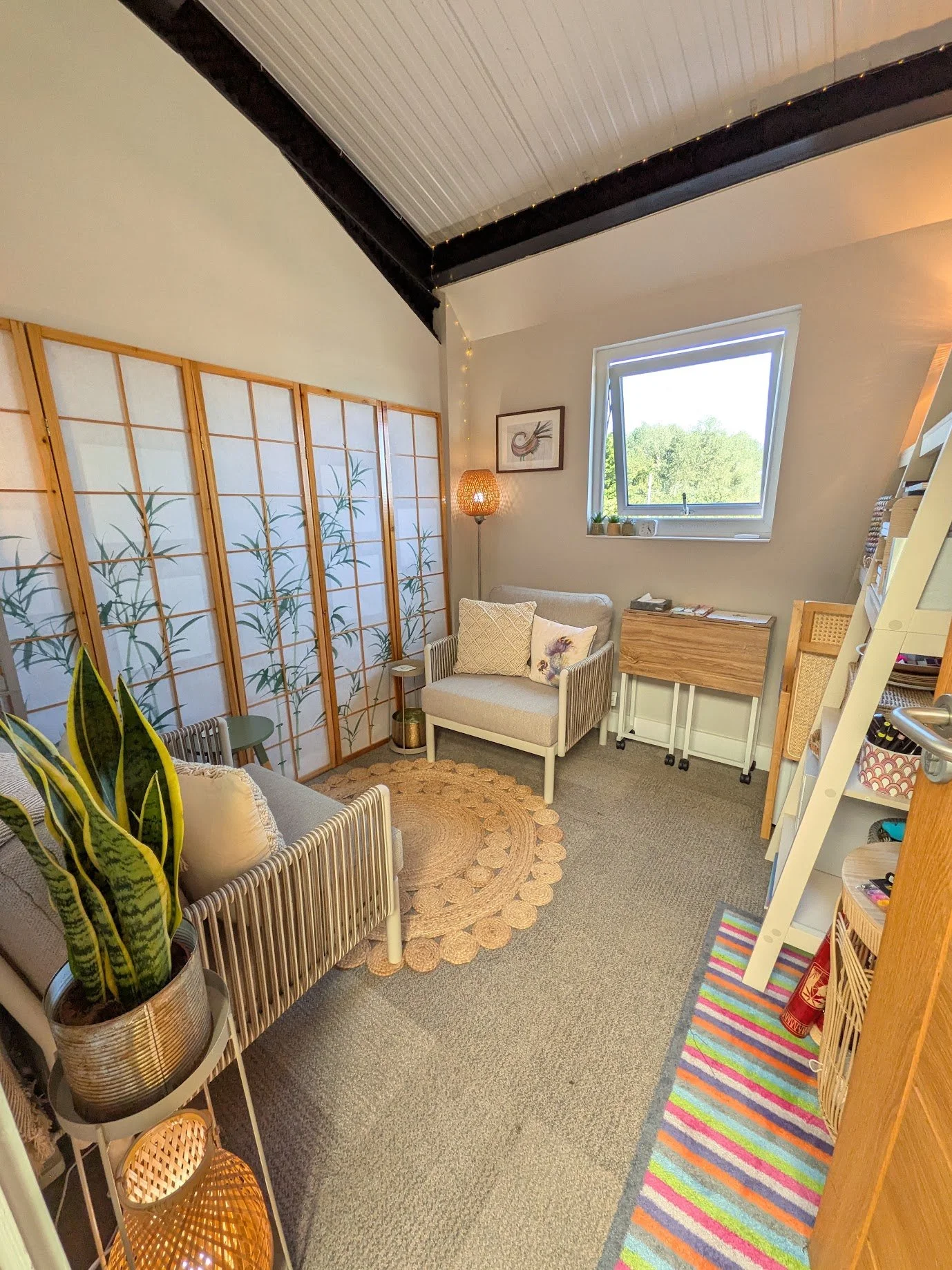 A room with a small sofa and armchair, a wooden cabinet, plants, a decorative screen with a botanical pattern, a window showing greenery outside, a round woven rug, and a colorful striped runner. There is a tall floor lamp next to the armchair and a ladder/bookcase on the right side.