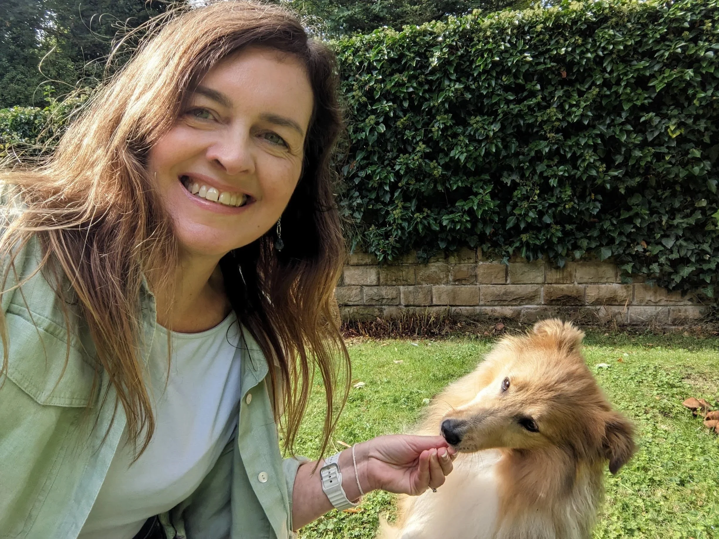 Therapist, Davina Hanlon, with long brown hair wearing a green shirt, smiling while feeding Barney, a brown and white dog outdoors in a garden with green bushes and a brick wall in the background.