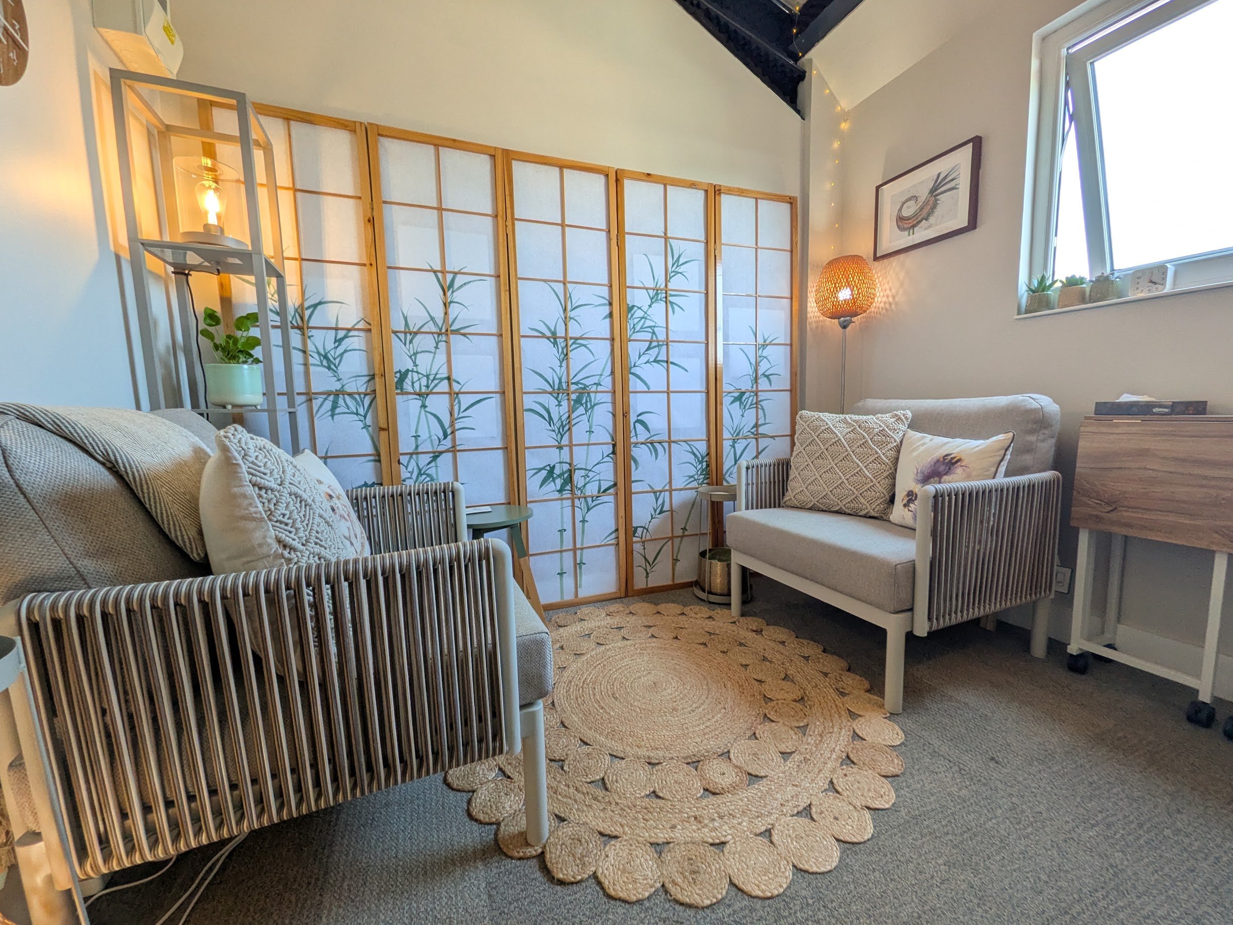 Therapy room with beige chairs, patterned pillows, woven round rug, green plant, folding room divider with bamboo design, small side table, window with potted plants, framed artwork, floor lamp, and string lights under a sloped ceiling.