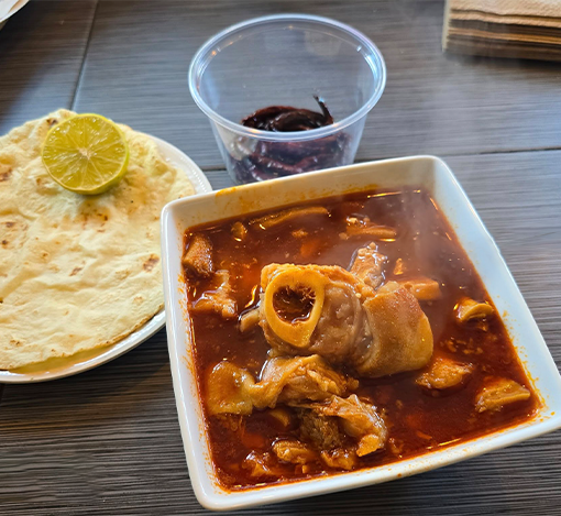 A plate of mashed potatoes with a lemon slice, a bowl of spicy stew with chicken and vegetables, and a clear plastic cup with a dark red condiment or jelly on a wooden table.