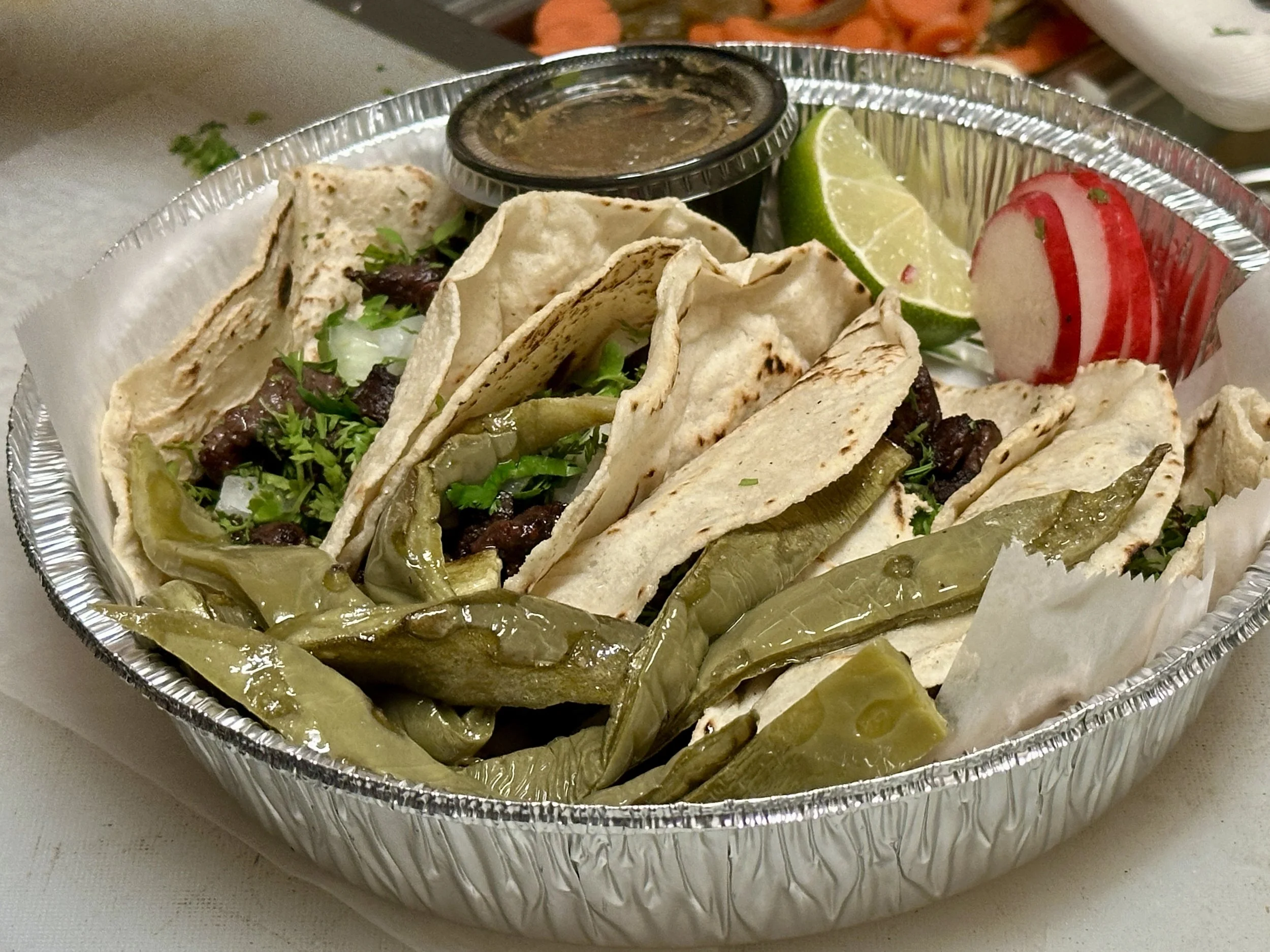 Aluminum tray containing tacos with beef, lettuce, and cilantro, served with lime, radish slices, pickled jalapeños, and a small container of sauce.