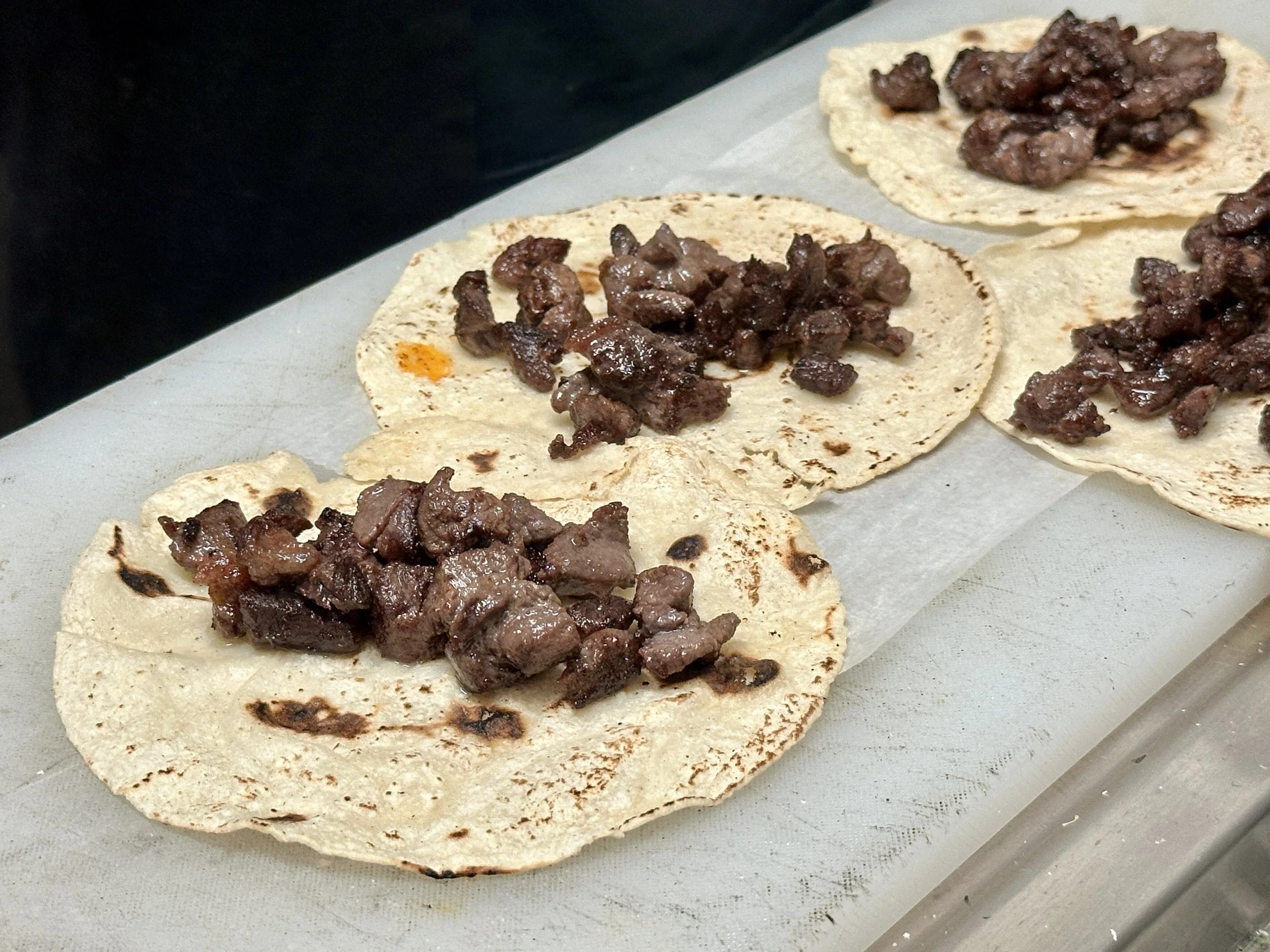 Tacos with grilled meat on corn tortillas placed on parchment paper.