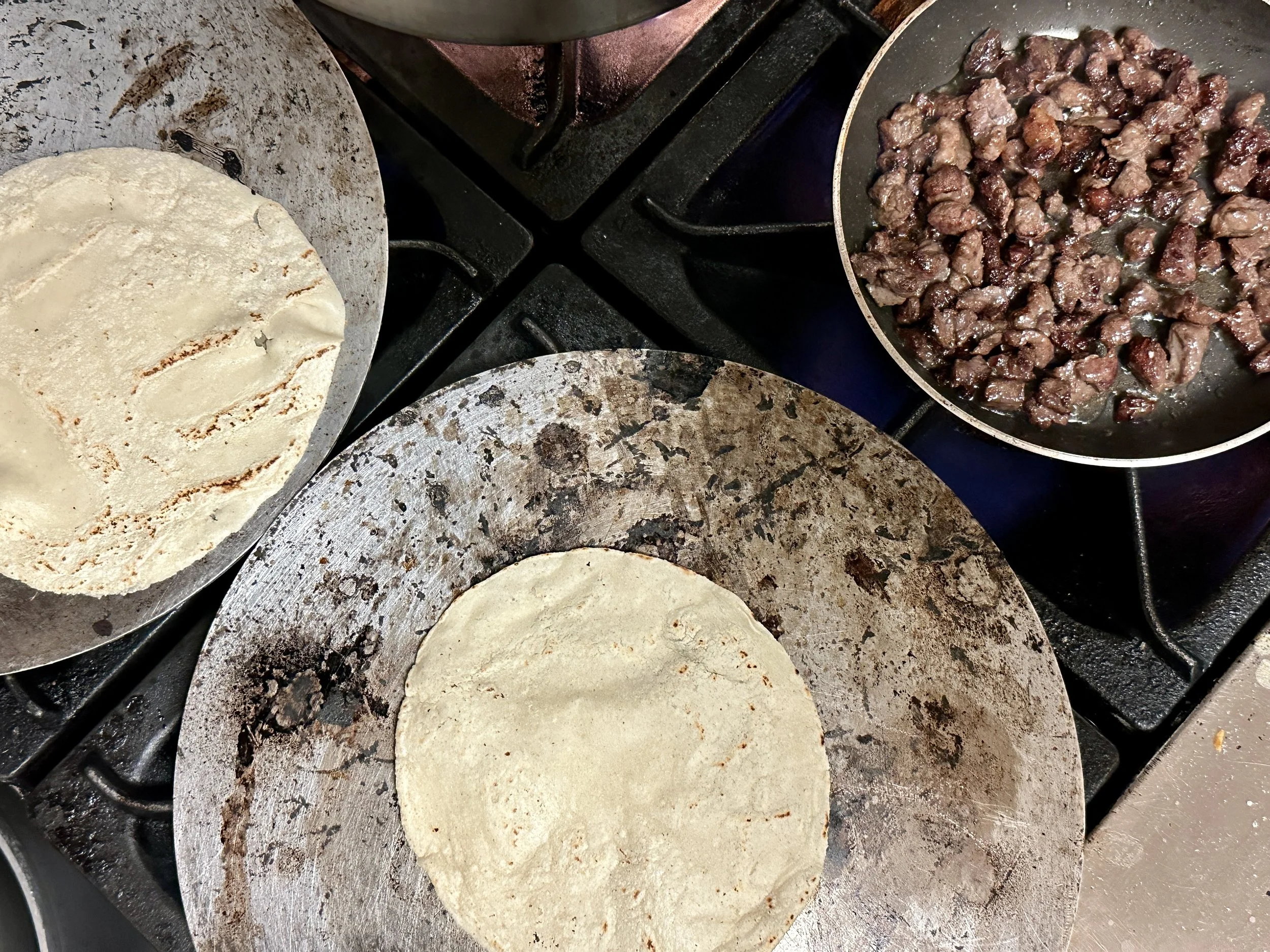Two tortillas on a round metal griddle, cooked to a golden brown, with a pan of cooked ground meat on a stove in the background.