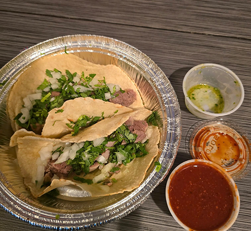 Aluminum tray holding two beef tacos with chopped onions and cilantro, with three small containers of green salsa, red salsa, and orange sauce on a wooden table.