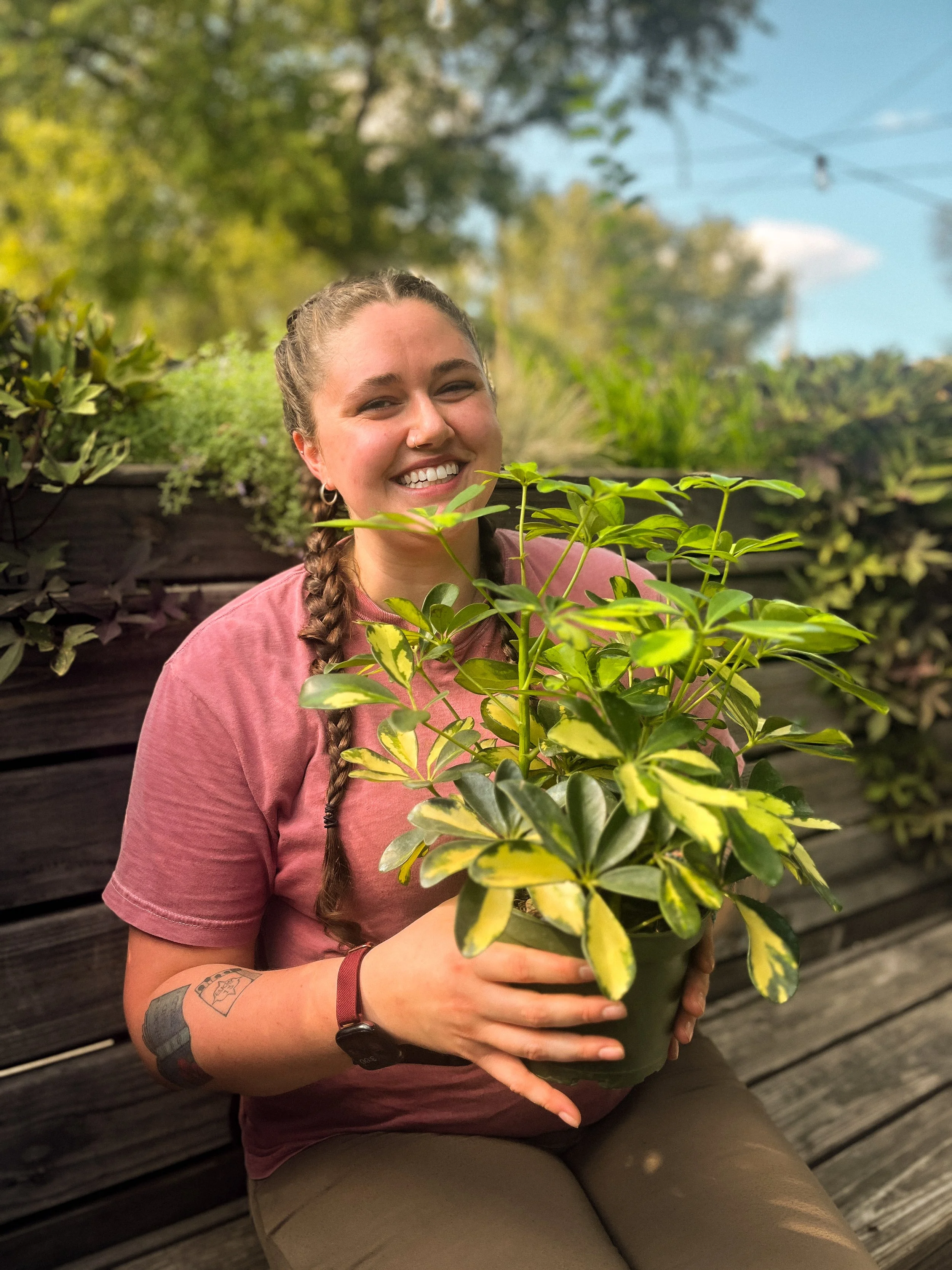 A smiling woman with braided hair, wearing a pinkish T-shirt and khaki pants, holding a potted plant with green and yellow variegated leaves while sitting outdoors on a wooden bench with greenery in the background.