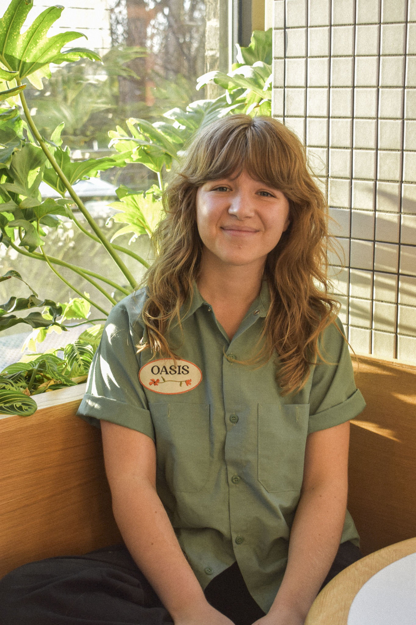 Young woman with shoulder-length red hair smiling outdoors on a rooftop with plants, flowers, and a city skyline in the background.