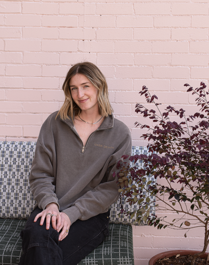 A woman with shoulder-length light brown hair, wearing a brown sweatshirt with 'OASIS DESIGN' and a line drawing of a flower, sitting indoors near a window with a tiled wall behind her and a large green plant nearby.