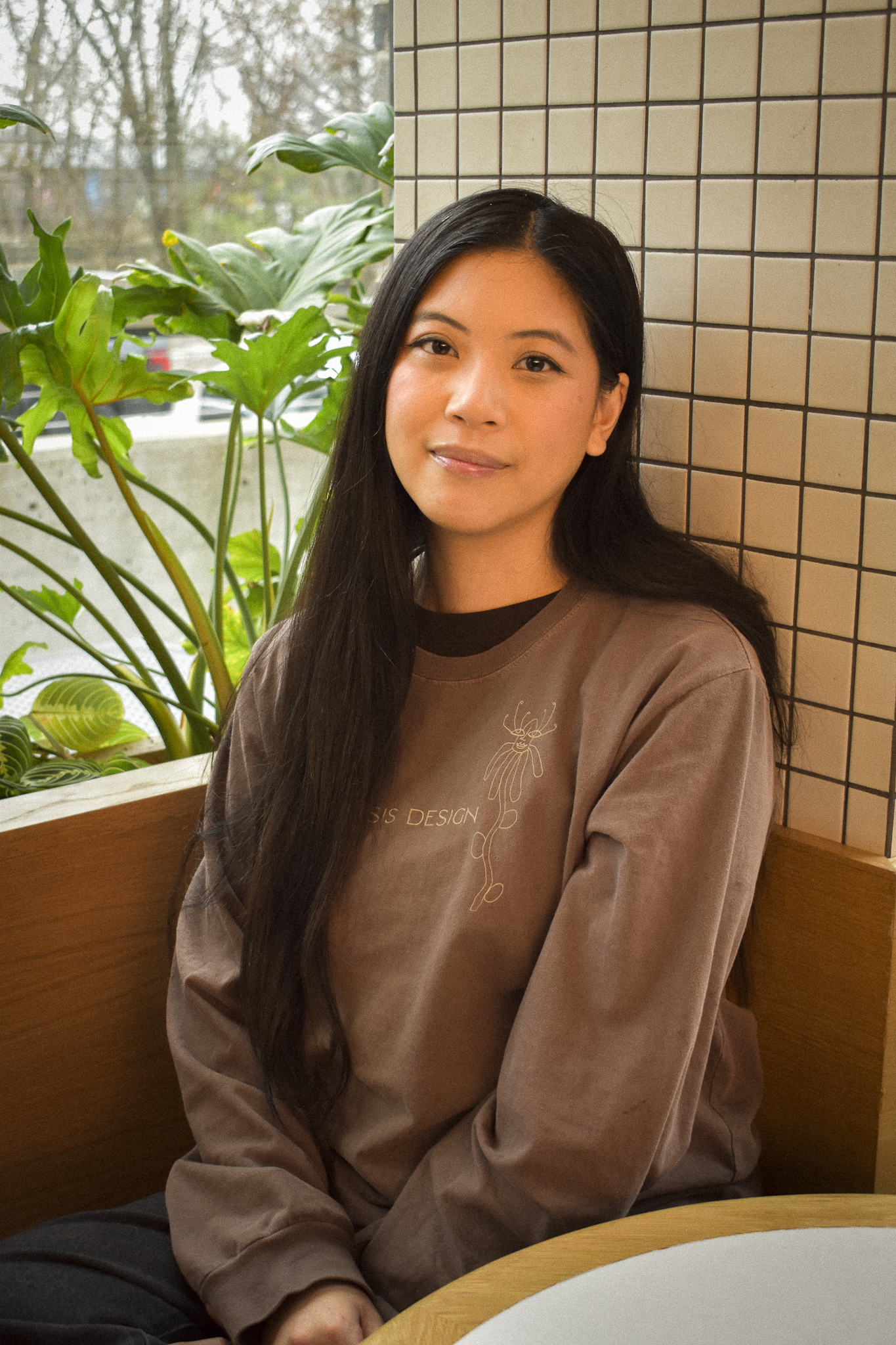 A young woman with long dark hair sitting on a wooden bench inside, behind her are large green plants and a window with trees outside.