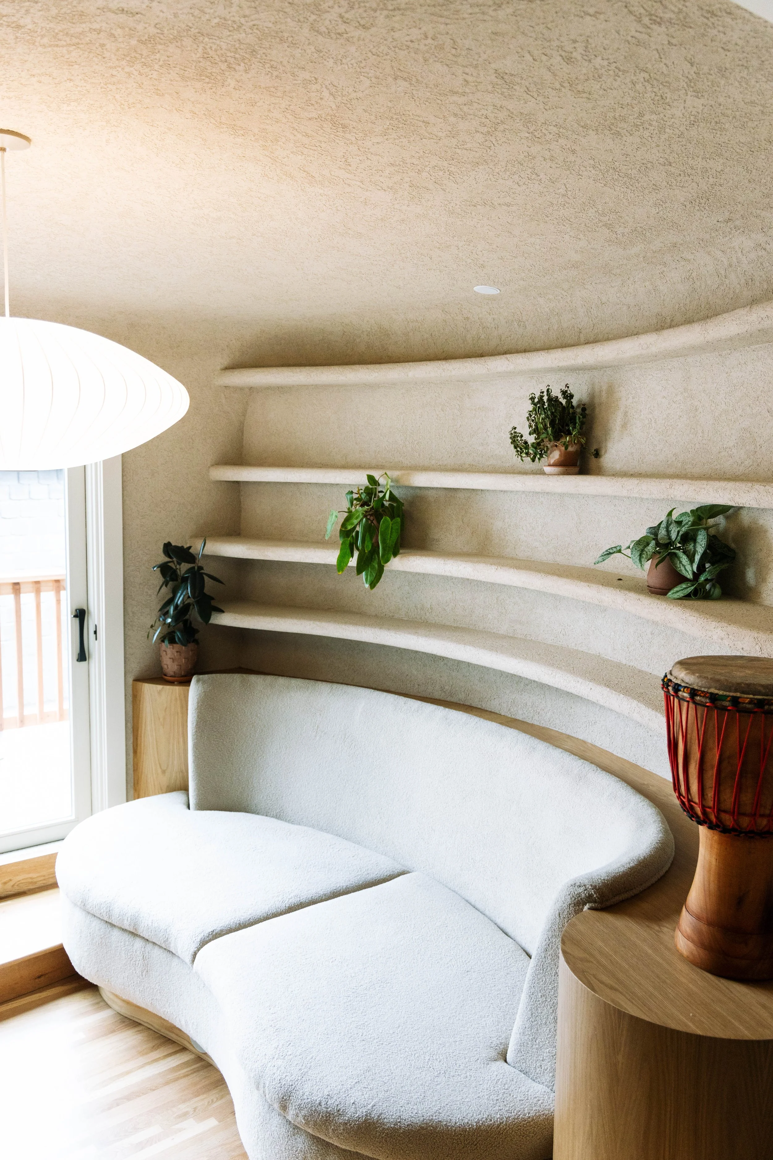 Interior of a living space with a curved white sofa, wooden side table, decorative drums, and built-in wall shelves with potted plants near a sliding glass door.