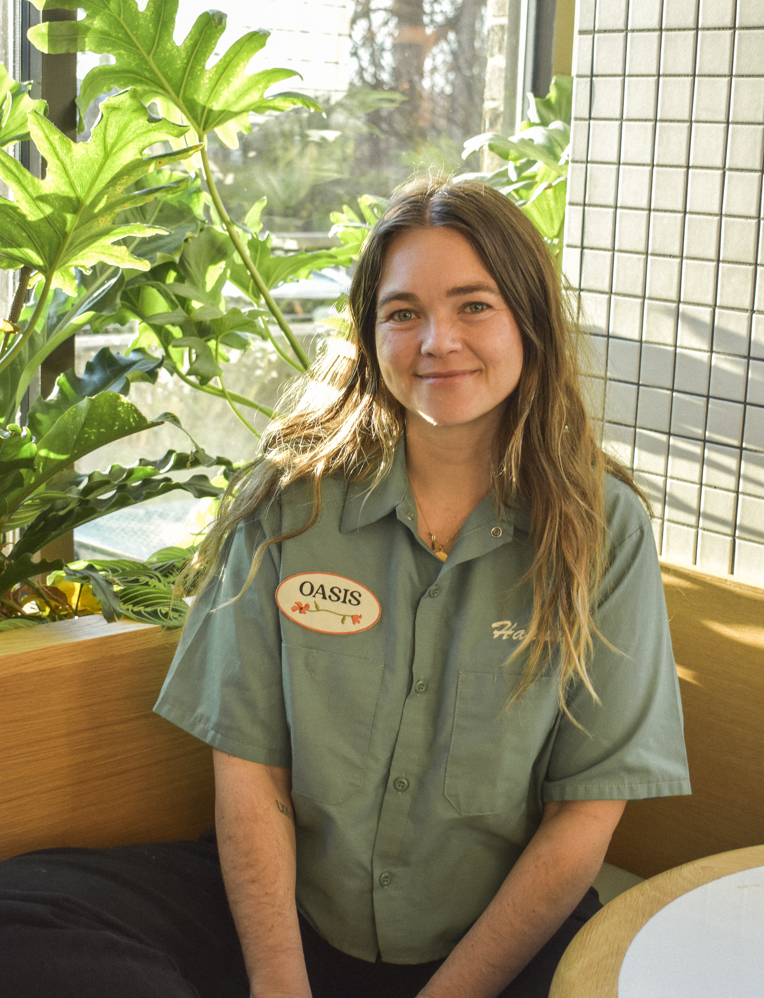 A young woman with long wavy hair wearing a gray t-shirt, black pants, and a cap standing outdoors in front of a decorative concrete block wall surrounded by potted plants.