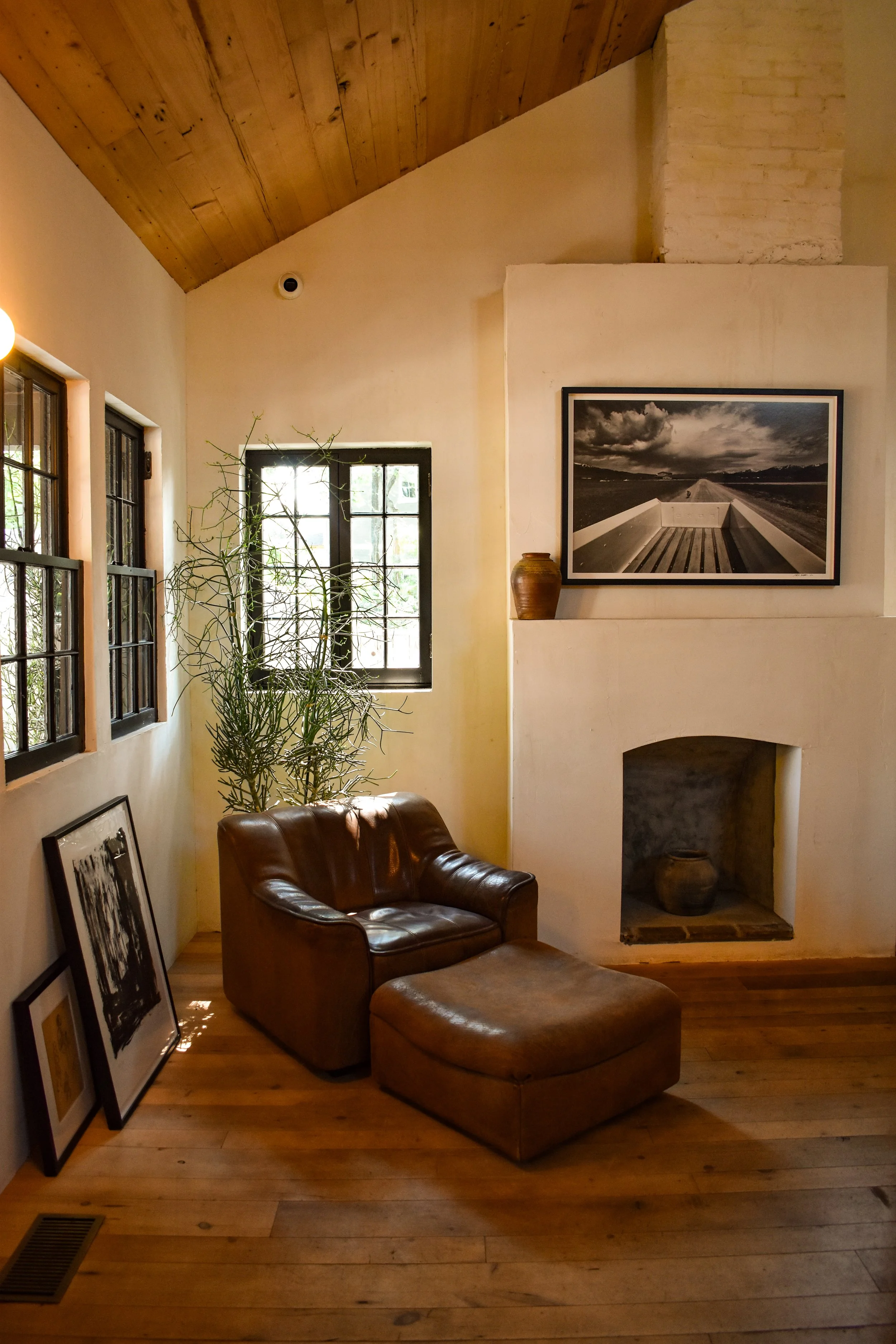 Cozy living room corner with a leather armchair and ottoman, framed art on the wall, tall plant by the window, and wooden ceiling and floor.