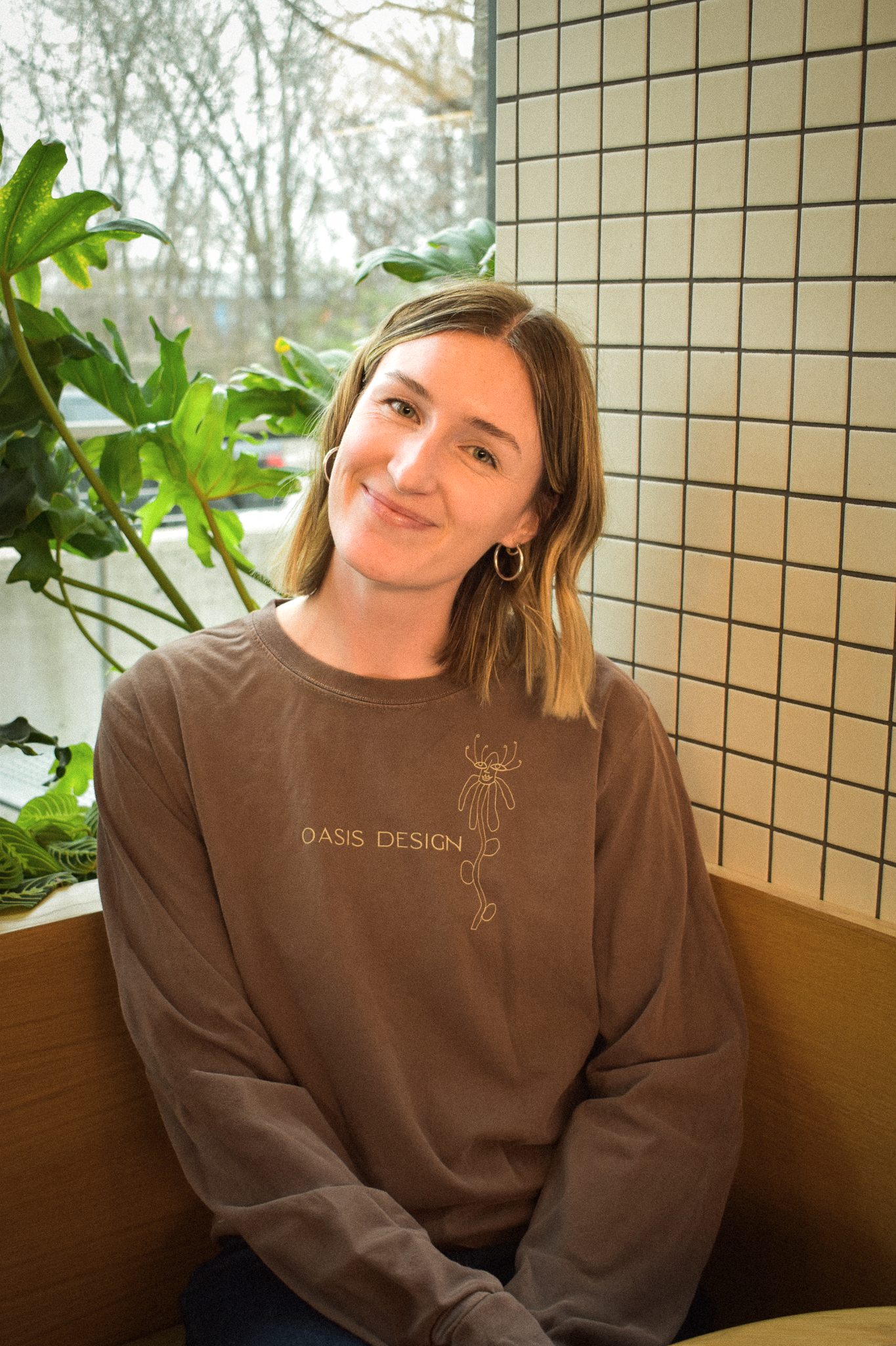 A woman with shoulder-length light brown hair, wearing a brown sweatshirt with 'OASIS DESIGN' and a line drawing of a flower, sitting indoors near a window with a tiled wall behind her and a large green plant nearby.