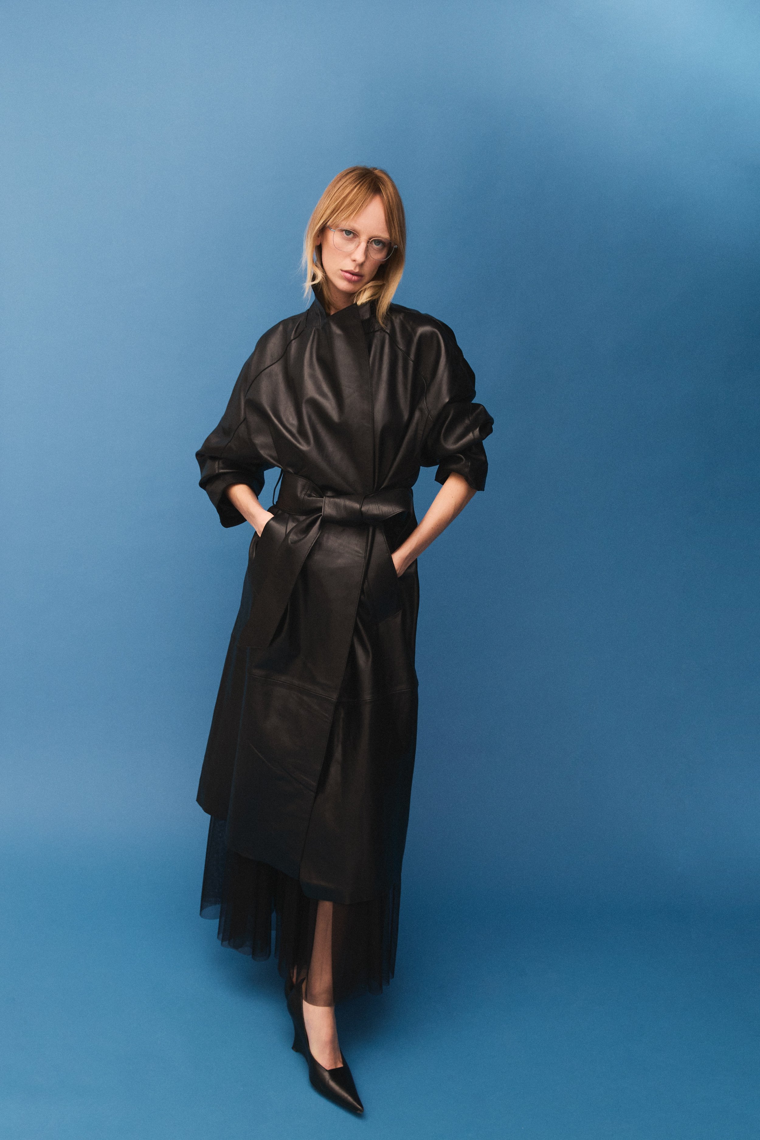 A woman with blonde hair and glasses wearing a black leather dress with a sheer black skirt standing against a blue background.