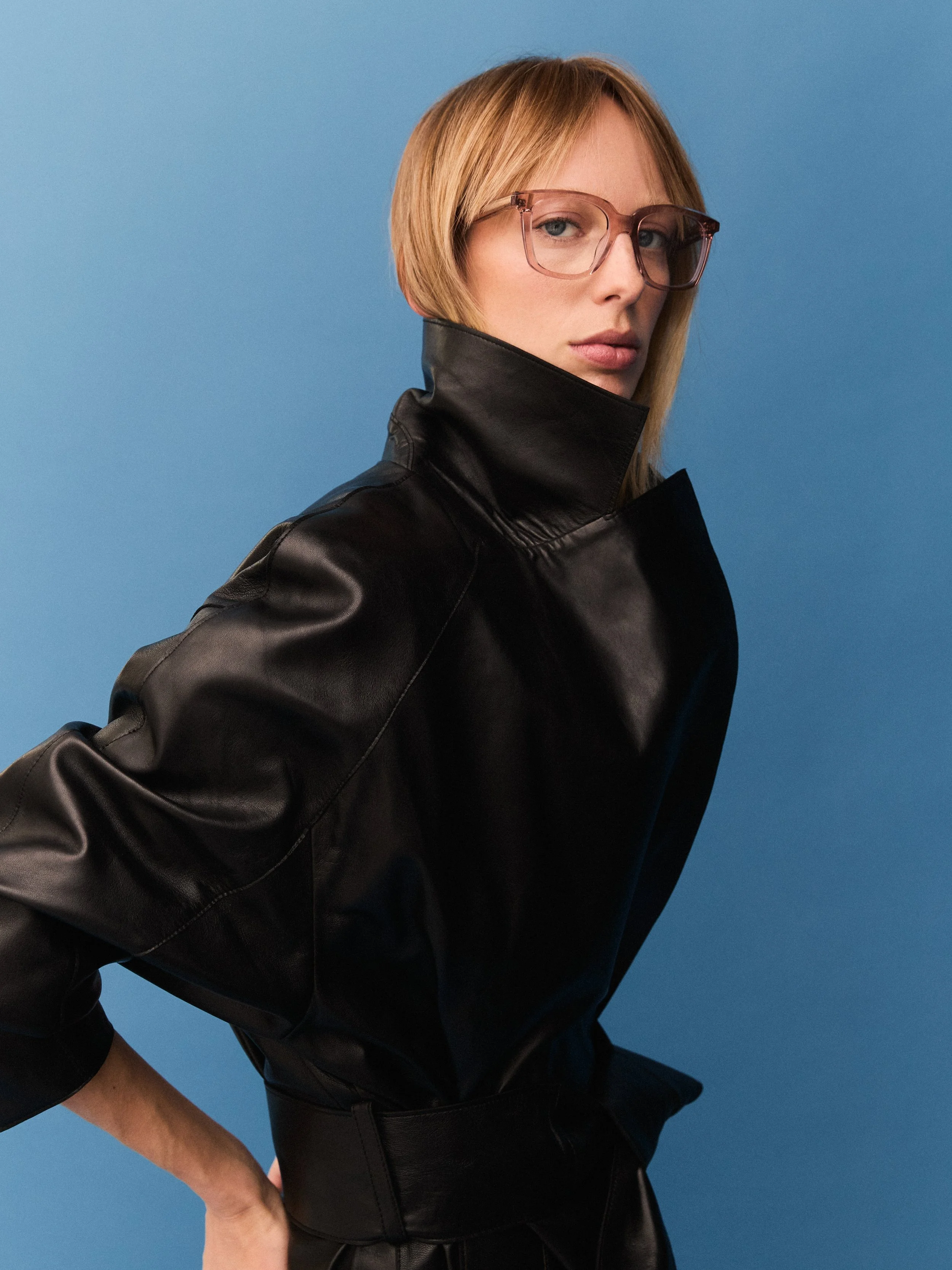 A woman with blonde hair wearing glasses and a black high-collared leather jacket posing against a blue background.