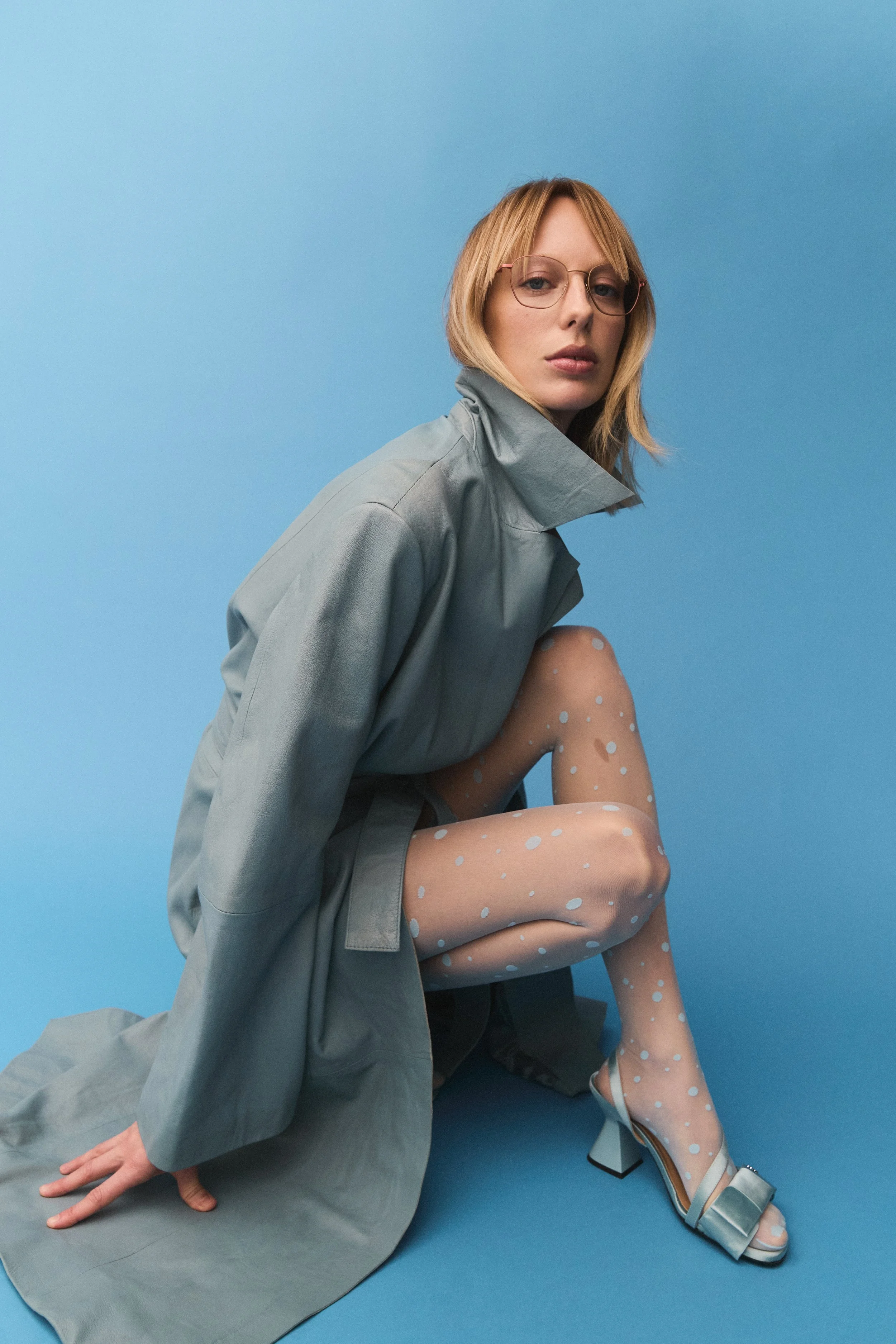 A woman with blonde hair, glasses, and wearing a gray trench coat, patterned sheer tights with white dots, and light blue high-heeled sandals, posing against a blue background.