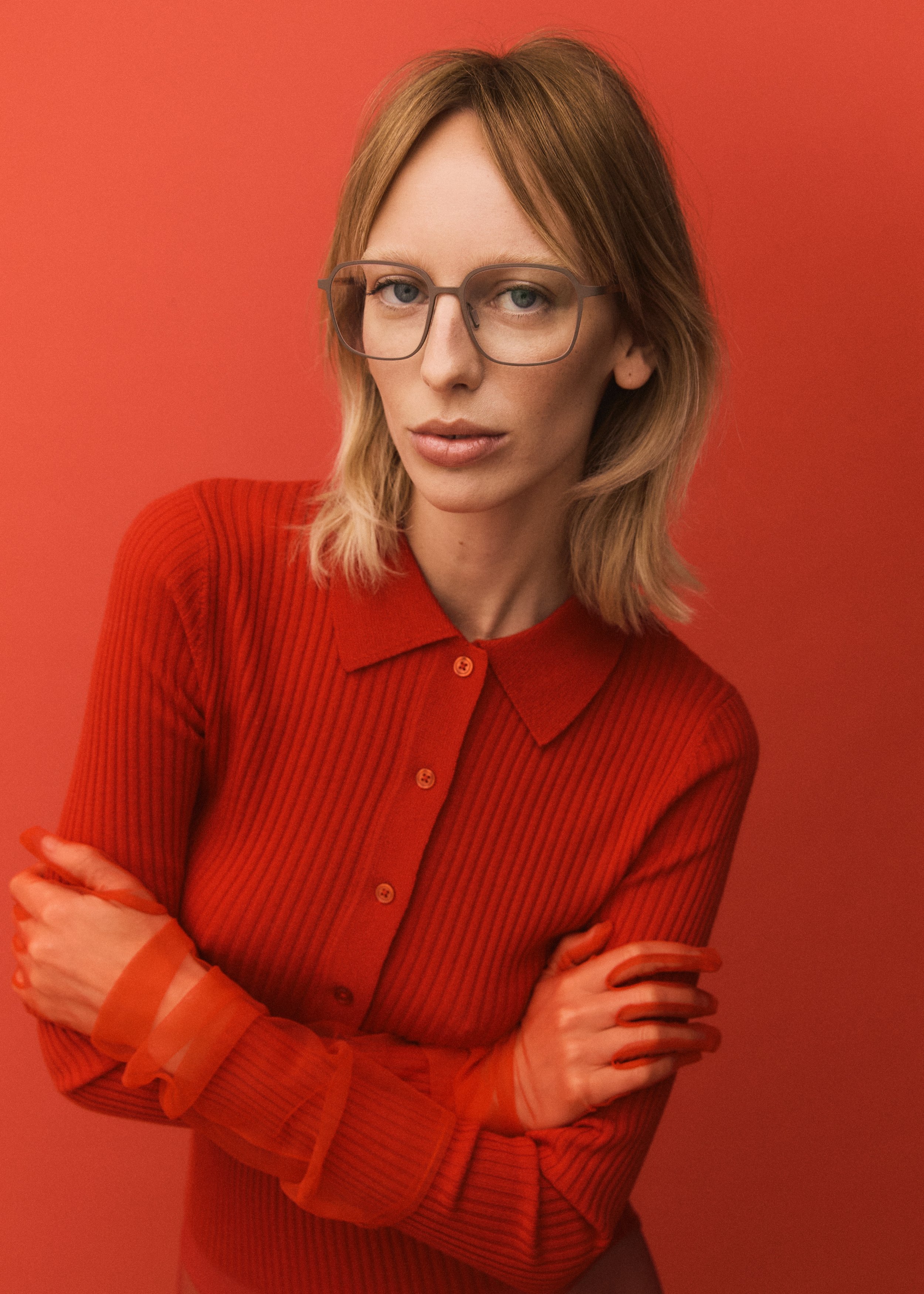 A woman with blonde hair and glasses wearing a red ribbed long-sleeve shirt against a red background.
