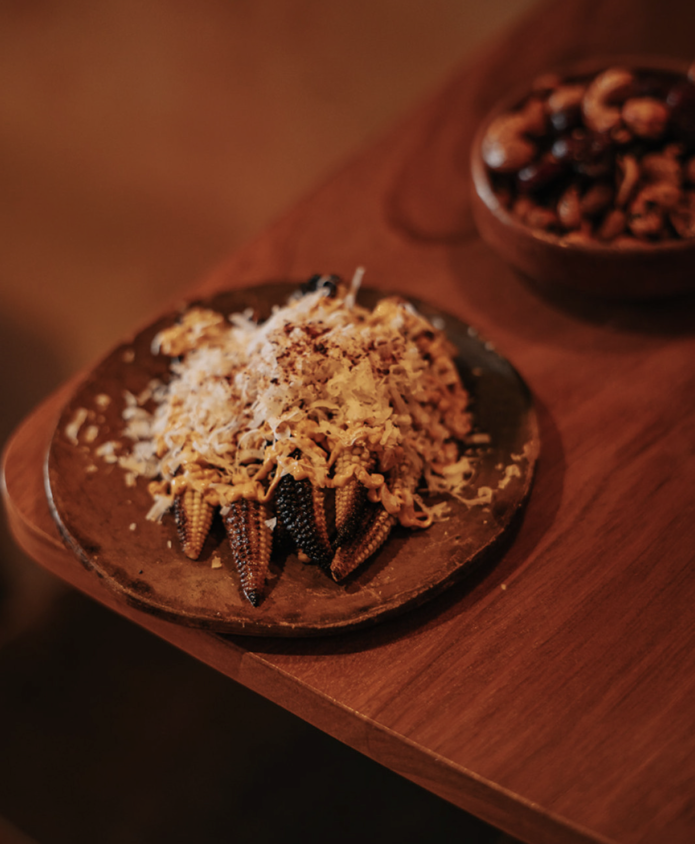 Two small round decorative dishes on a wooden surface, one with a chocolate dessert topped with shredded coconut and young corn cobs, the other with chocolate-covered cookies or nuts.