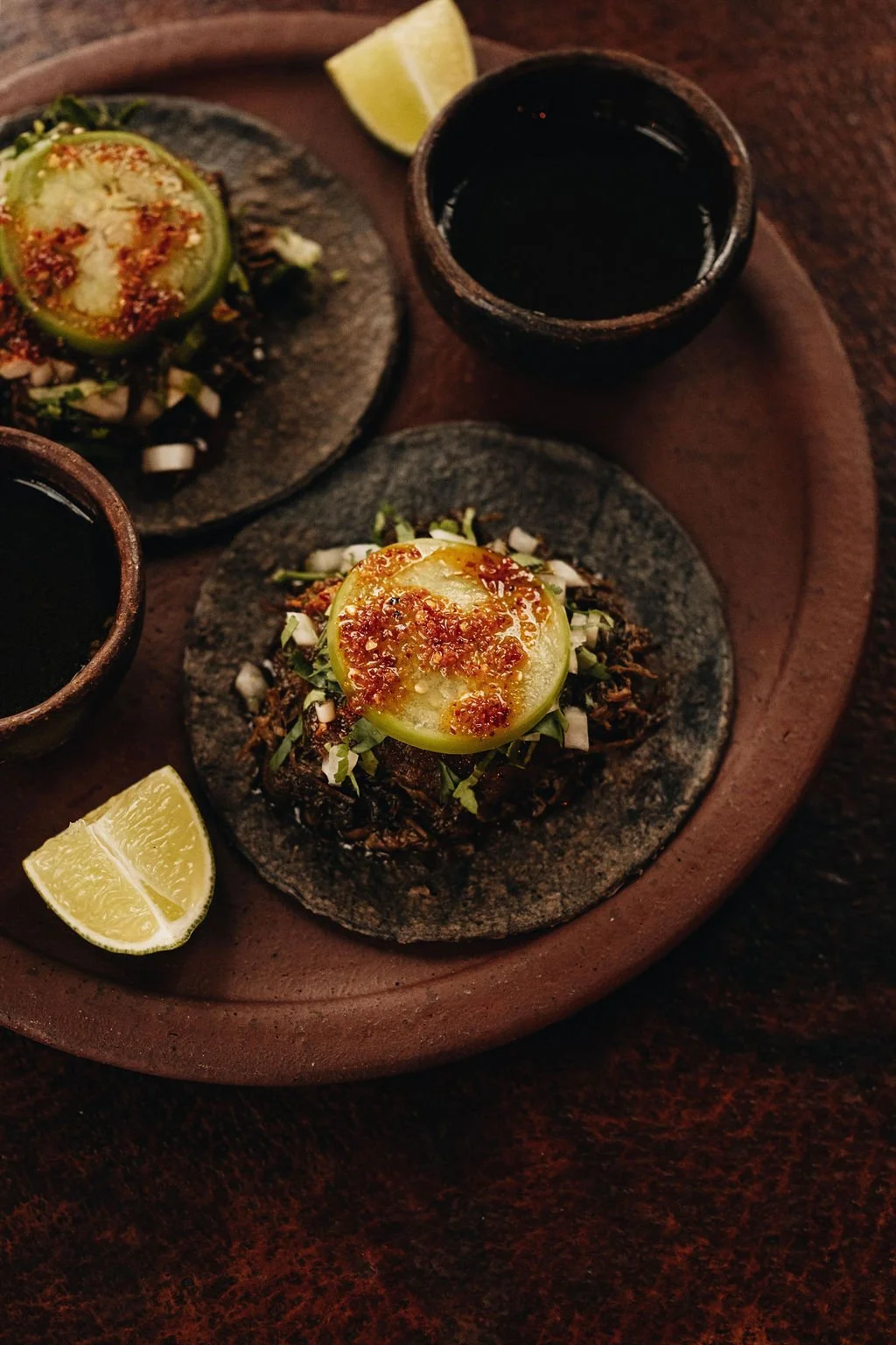 Two tostadas topped with sliced avocado, chopped onions, cilantro, and spicy sauce, served with lime wedges and two cups of dark beverage on a wooden tray.