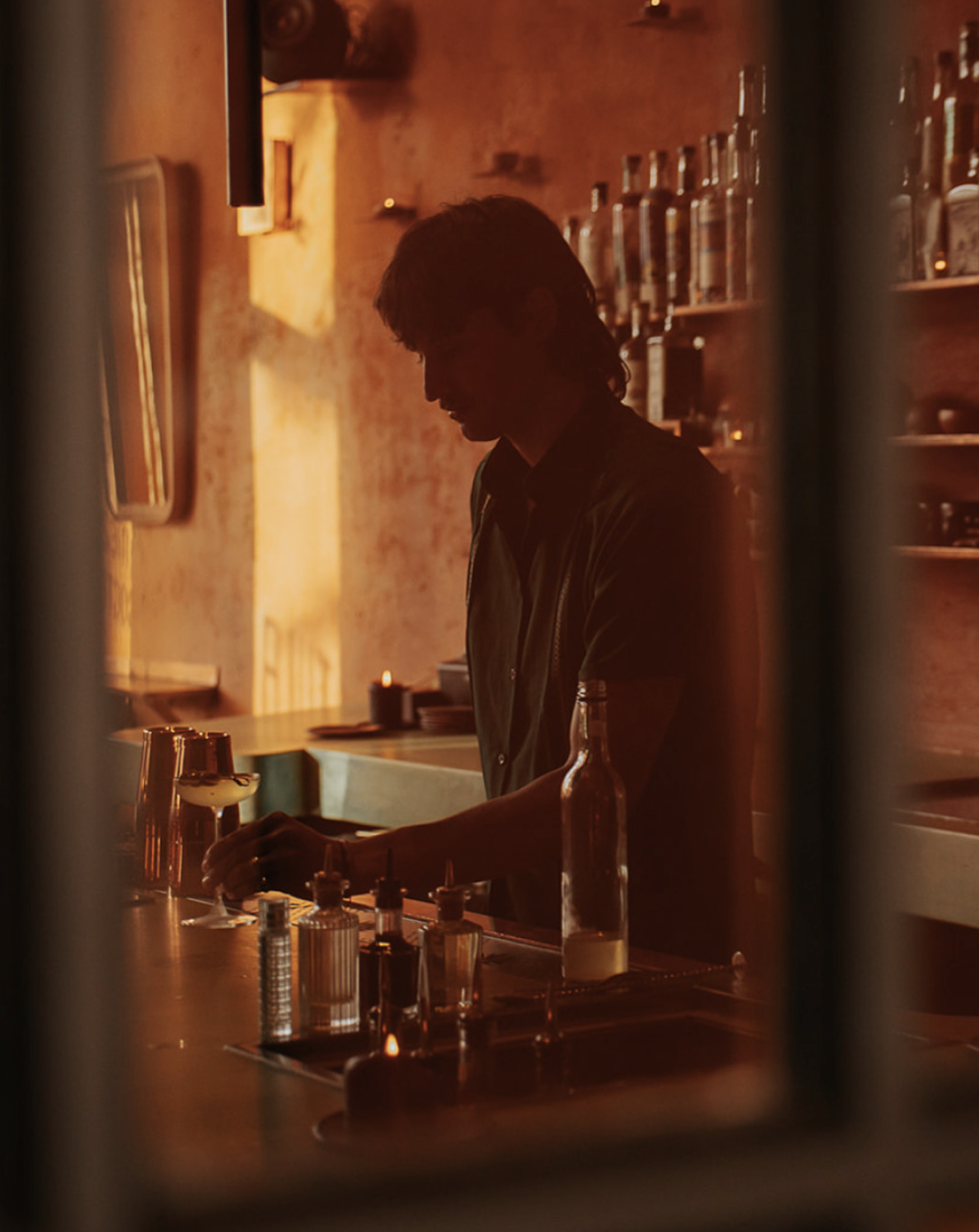Silhouette of a person, likely a bartender, preparing drinks behind a bar in a dimly lit, warm-toned environment with bottles on shelves in the background.