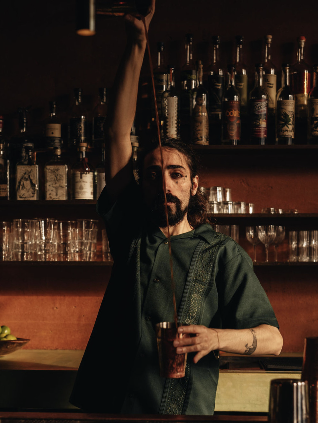 A man with long hair and a beard pouring a drink from a cocktail shaker into a glass at a bar with bottles on shelves behind him.