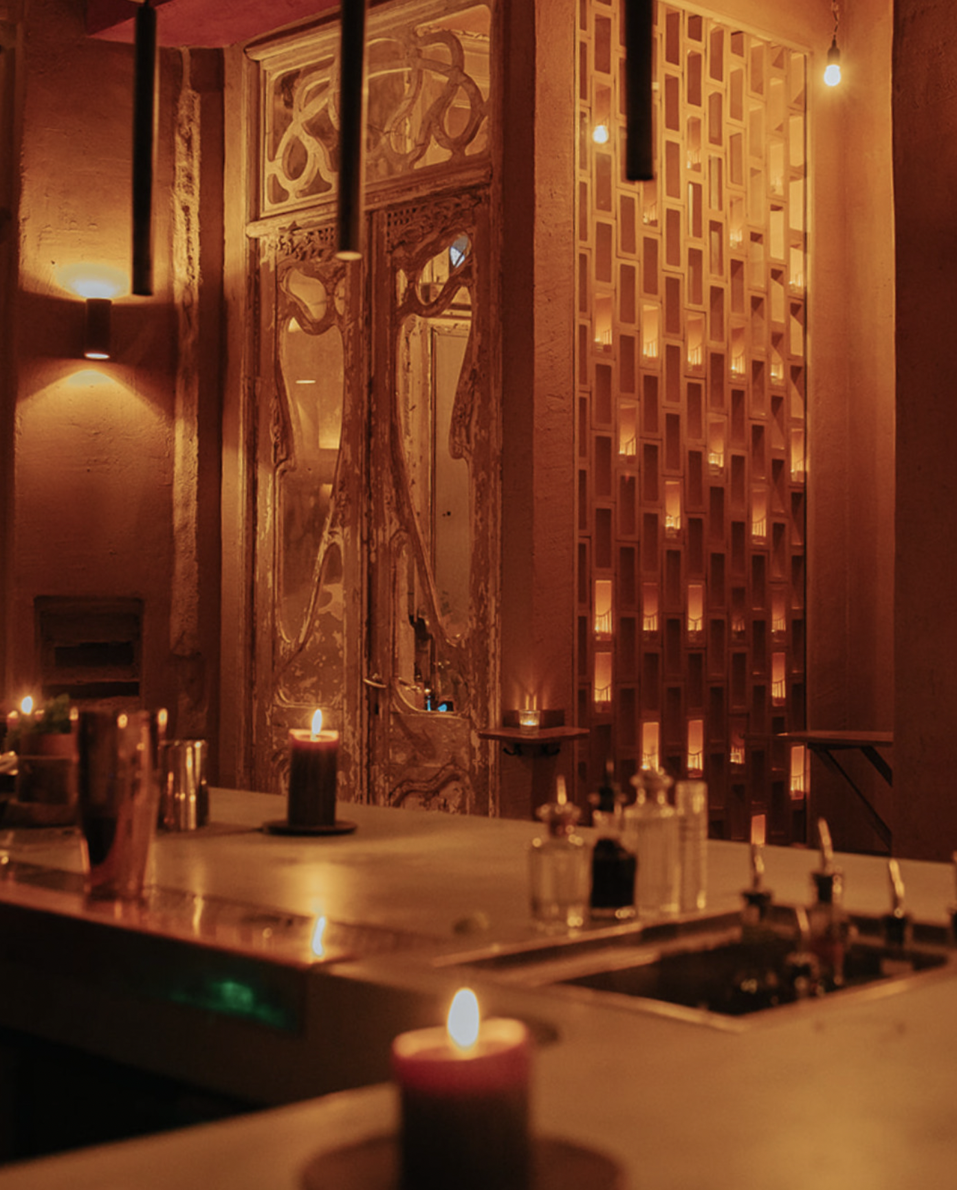 Dimly lit interior of a restaurant with candles on tables, warm amber lighting, a decorative wooden lattice wall, wooden framed mirrors, and exposed light bulbs.