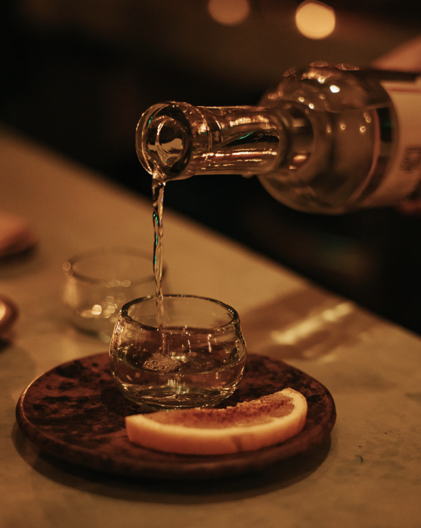 A bottle of clear liquor is pouring into a glass from its neck, with a slice of lemon on a wooden plate in the foreground, set against a warm, dimly lit background.