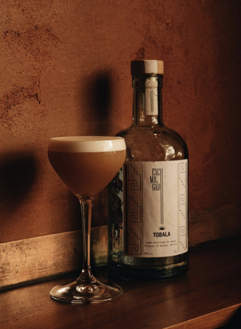 A cocktail in a coupe glass with a frothy top sits on a wooden surface next to a bottle labeled 'TOBALA' with ornate design.