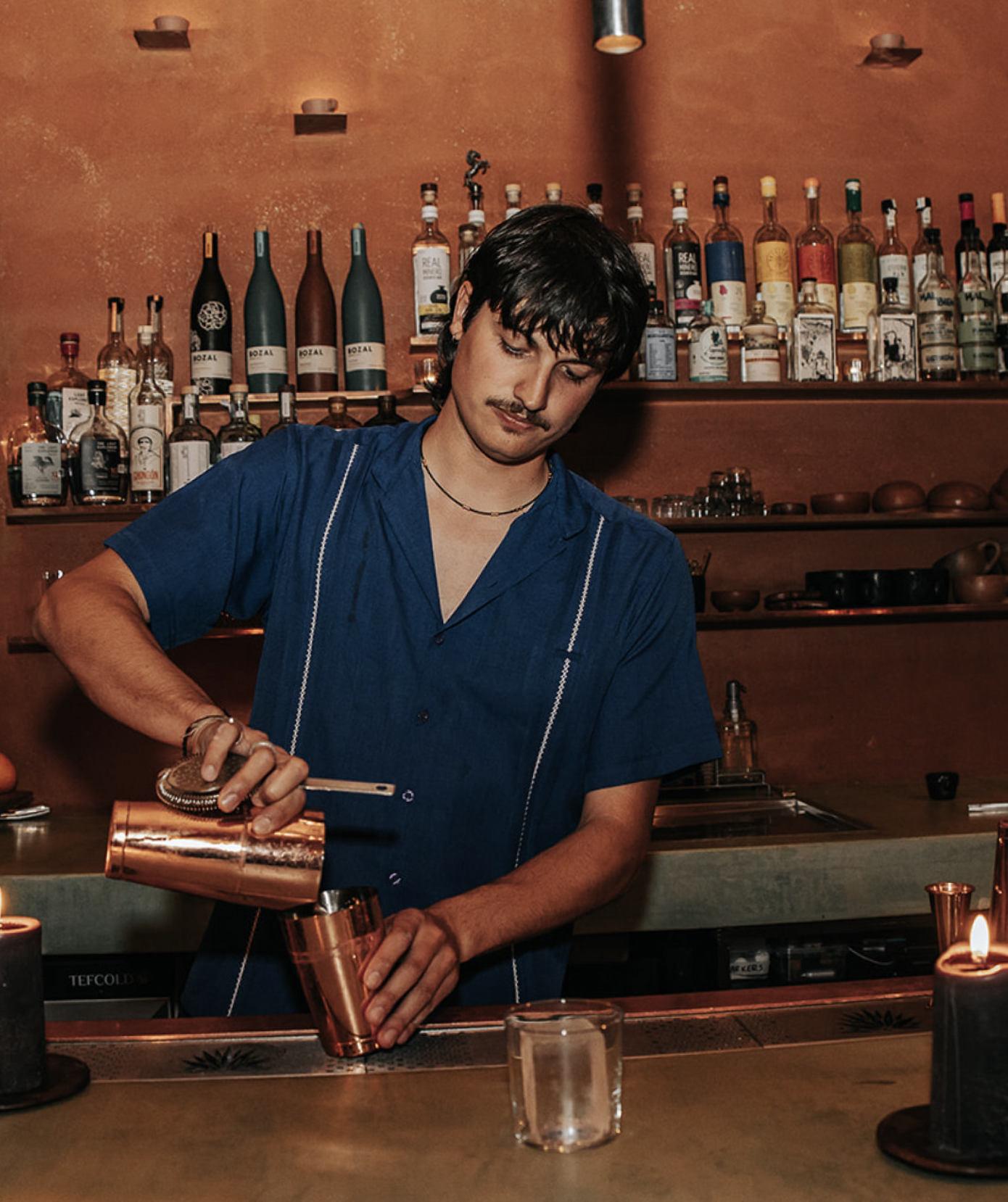 A bartender with dark hair and a mustache pouring a drink from a copper shaker into a copper cup at a bar with shelves of alcohol bottles in the background.