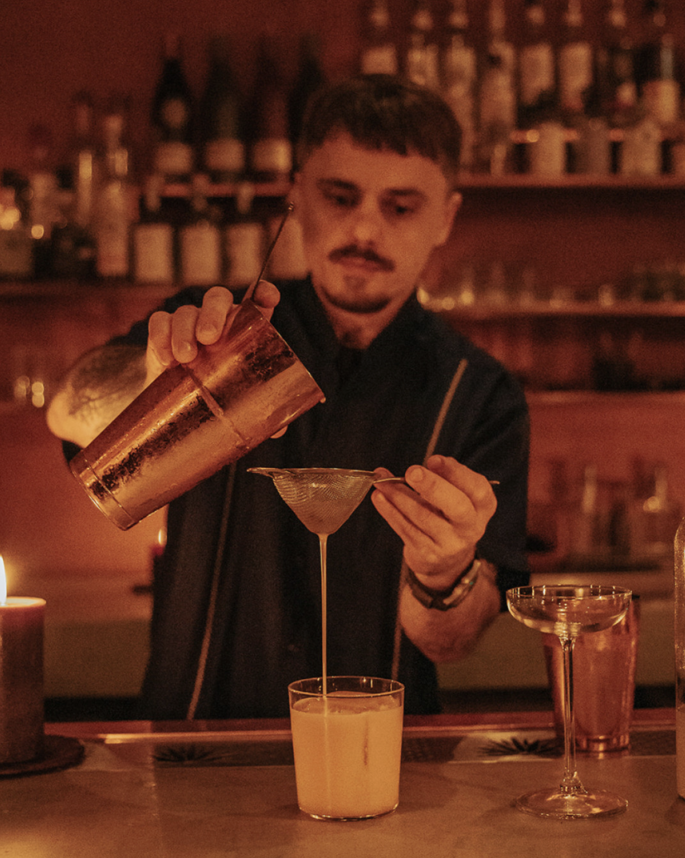 A bartender pouring a yellow cocktail from a shaker into a tall glass through a strainer in a dimly lit bar with shelves of bottles in the background.