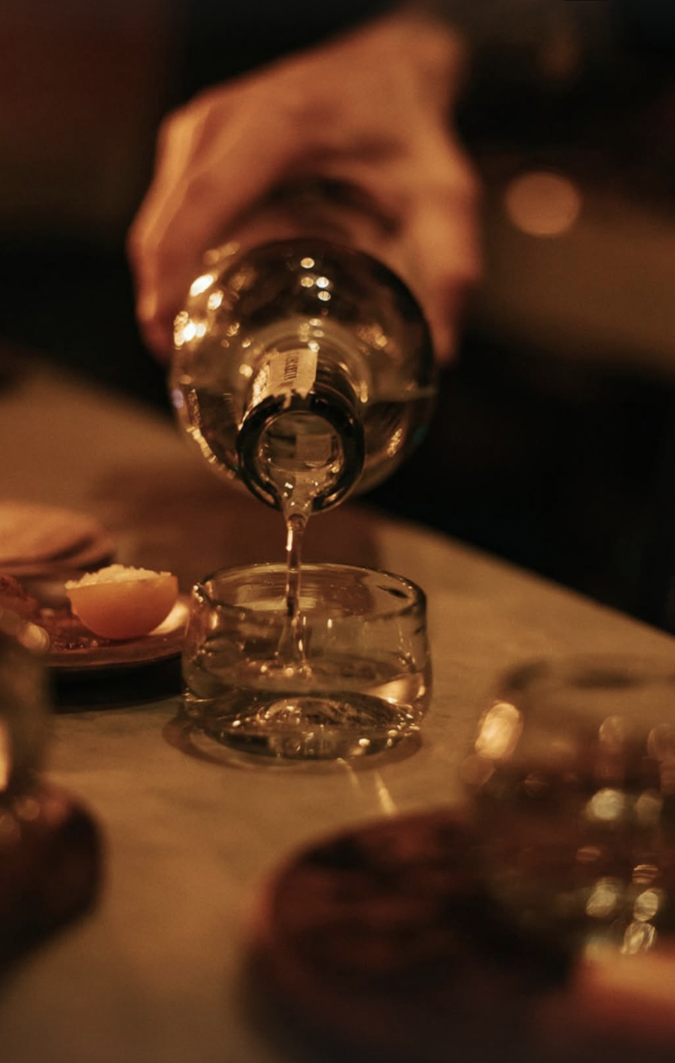 A hand pouring water from a bottle into a glass on a table with various dishes and dim lighting.