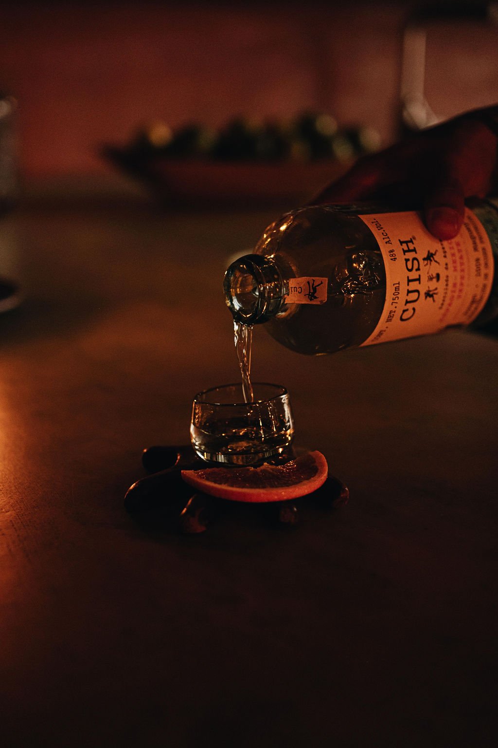 A hand pouring liquid from a bottle labeled 'CUSH' into a small glass placed on a dark surface. The scene is dimly lit with a warm glow, and a slice of citrus fruit is positioned beneath the glass.