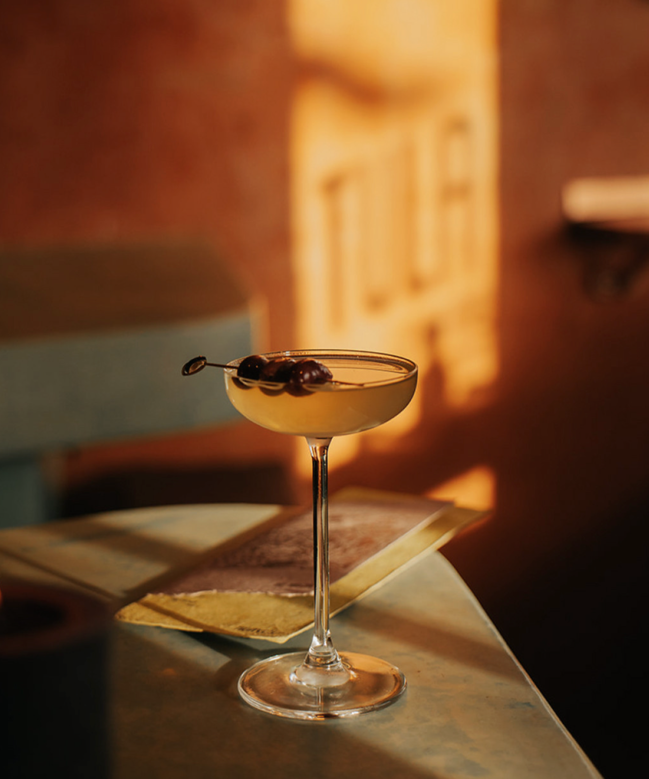 A cocktail glass with dark cherries garnishing the drink, placed on a table with open books nearby, in a dimly lit setting.