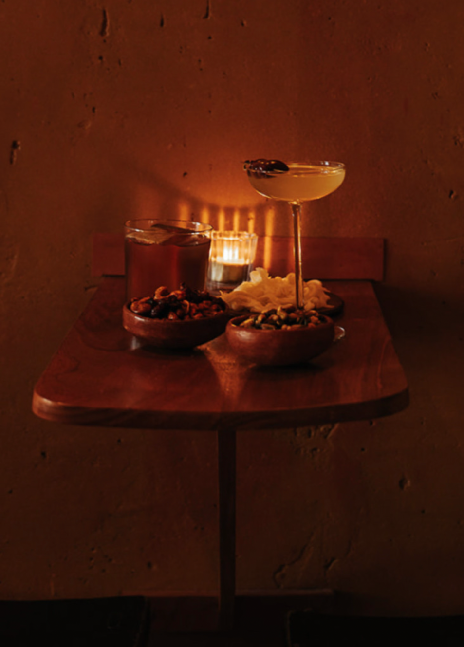 A dimly lit bar or lounge table setting with drinks, snacks, and a candle.
